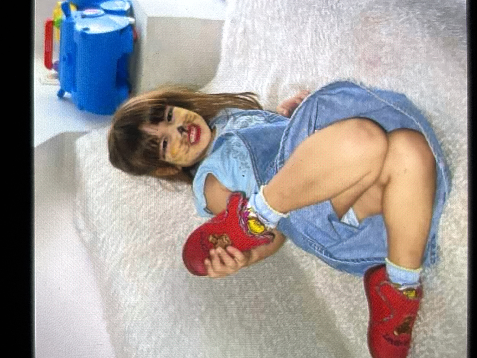 A young child with face paint resembling a cat is lying on a textured, white surface. The child wears a denim dress or overalls and bright red shoes with yellow accents. One of the child's hands is holding their foot, which is clad in one of the red shoes. In the background, a blue toy is visible. The lighting suggests an indoor setting. The child's expression is difficult to discern due to the face paint, but the overall scene appears playful. The location context is Burgas, Bulgaria. There is no visible text in the image.