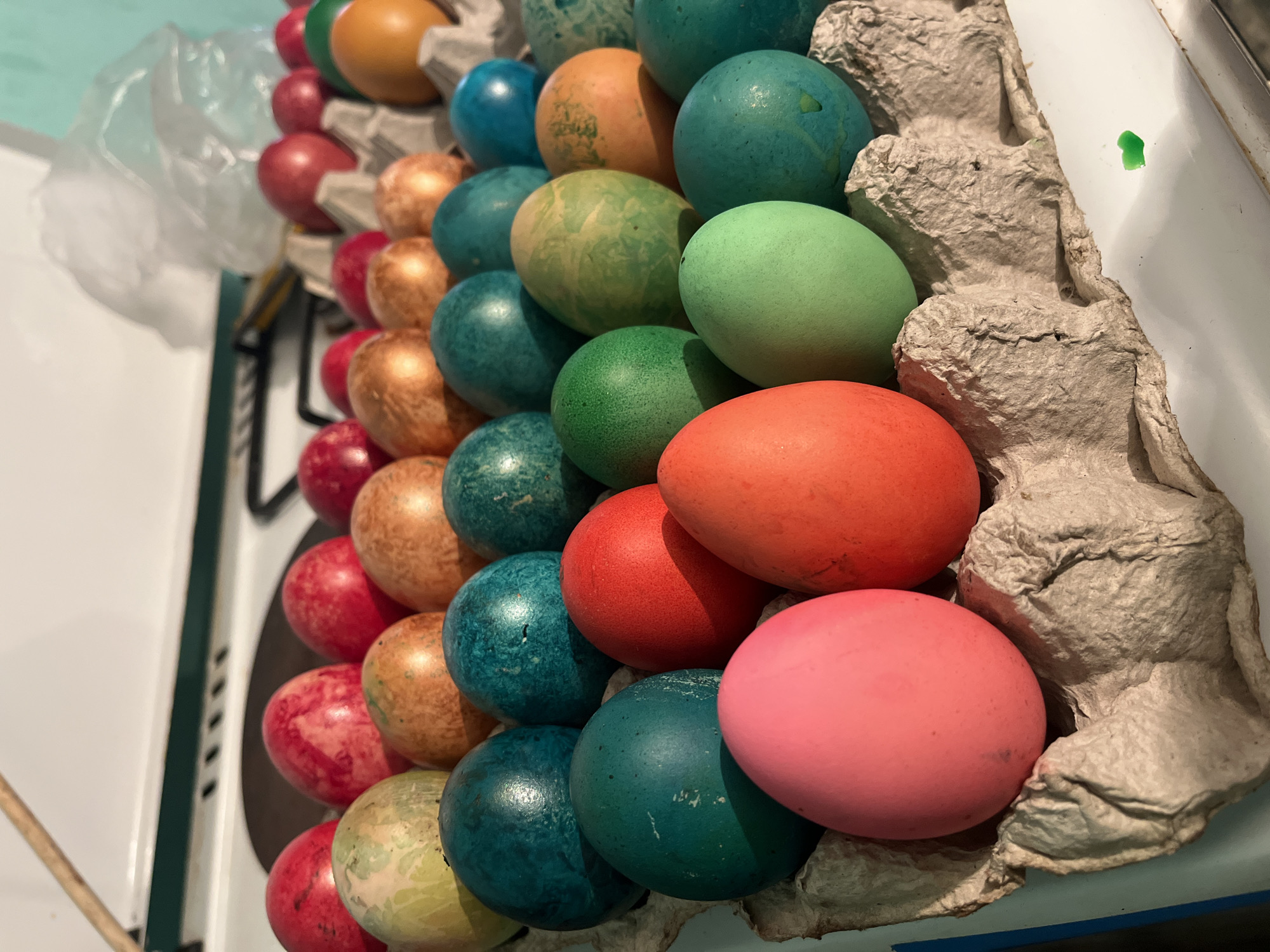 A close-up, high-angle shot shows a carton of dyed Easter eggs. The eggs, which are in rows, fill the carton. They are painted in various colors including red, gold, blue, green, orange and pink. The carton appears to be constructed of recycled paper. The background is a white stovetop. The location of the image is Lozarevo, Bulgaria, based on visual analysis and provided context. No people are visible.