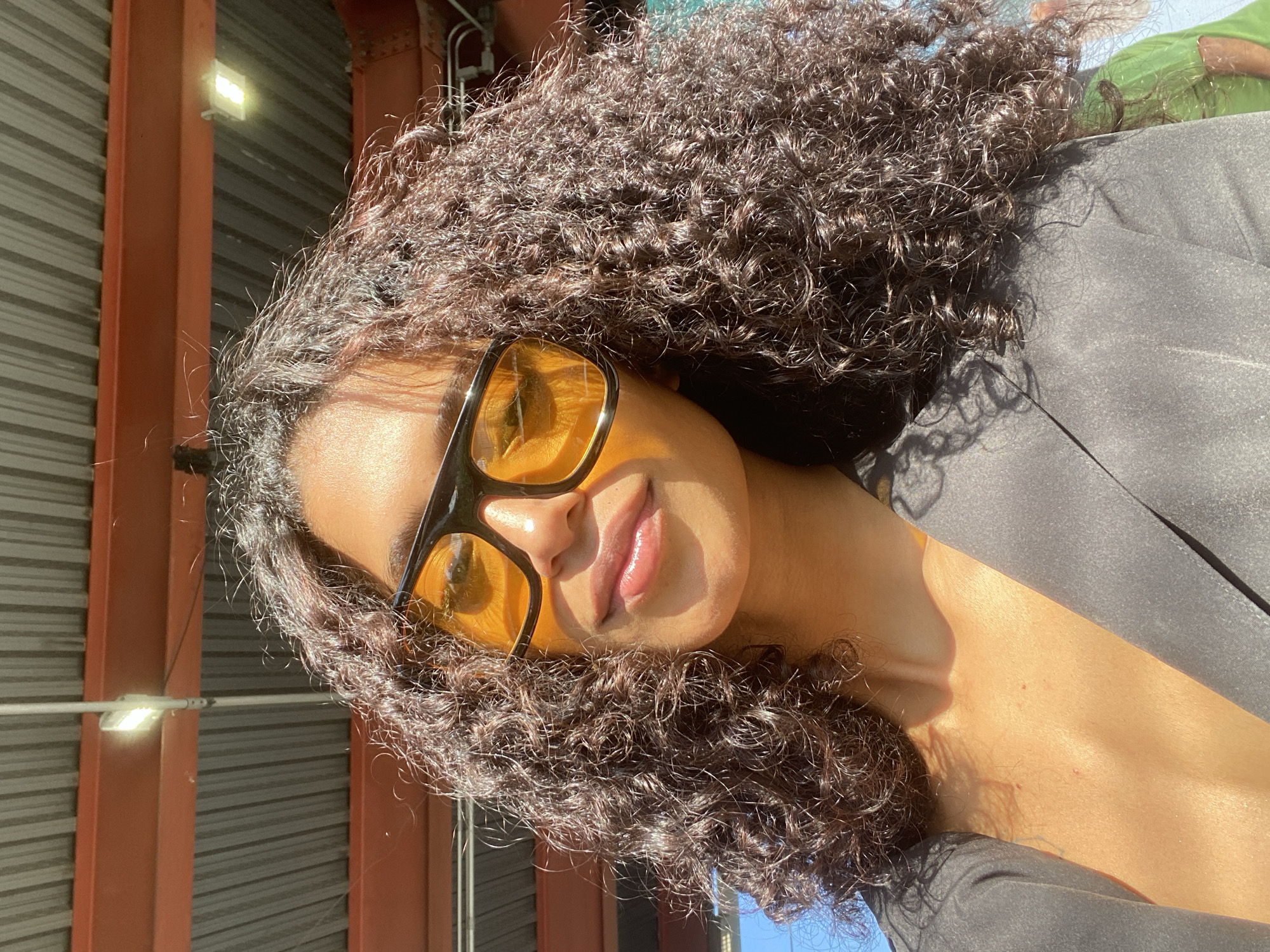 The image features a close-up portrait of a young woman with voluminous, dark brown curly hair, brightly illuminated by direct sunlight. She is wearing large, black-framed sunglasses with amber or yellow-tinted lenses, which obscure her eyes. Her lips are slightly parted in a subtle smile, with highlights from the sun. She is dressed in a dark, likely black, blazer or jacket, its fabric catching the light on her shoulder and chest. The strong sunlight creates warm highlights on her radiant skin and the intricate texture of her curls, while casting soft shadows.
The background reveals elements of an urban or industrial environment, including ribbed metal siding, robust orange structural beams, and a prominent bright LED light fixture attached to one of the beams. A small section of clear blue sky is visible, confirming a sunny day. The intense light and deep shadows suggest either early morning or late afternoon. The overall impression is one of modern style and confidence. No text is visible in the frame. The scene is set in São Paulo, Brazil.
