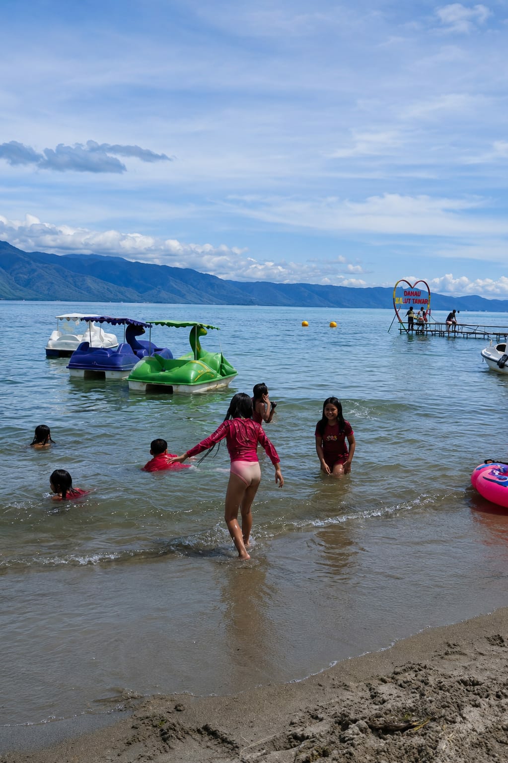 A group of children are swimming and playing in the shallow waters of a lake, near the shore. In the foreground, two girls in pink and maroon swimsuits are in the water, with one girl in the pink suit facing away from the camera, walking towards the deeper water. Several other children are visible in the water, some with their heads above the surface, also wearing swimsuits.
In the mid-ground, several colorful paddle boats are moored in the lake. Two are white and purple, and one is a bright green, shaped like a dinosaur. Yellow buoys are scattered in the water.
In the background, a wooden pier extends into the lake, with a large heart-shaped sign hanging above it. The sign has writing in Indonesian that appears to say "DANAU" and "ULUT TAWAR". Several people are standing on the pier, and a small white boat is docked to the right. Mountains line the horizon beyond the lake, under a bright blue sky with scattered clouds. The shoreline in the immediate foreground is a sandy beach with some debris. The overall scene depicts a recreational day at a lake in what appears to be a tropical or subtropical climate.