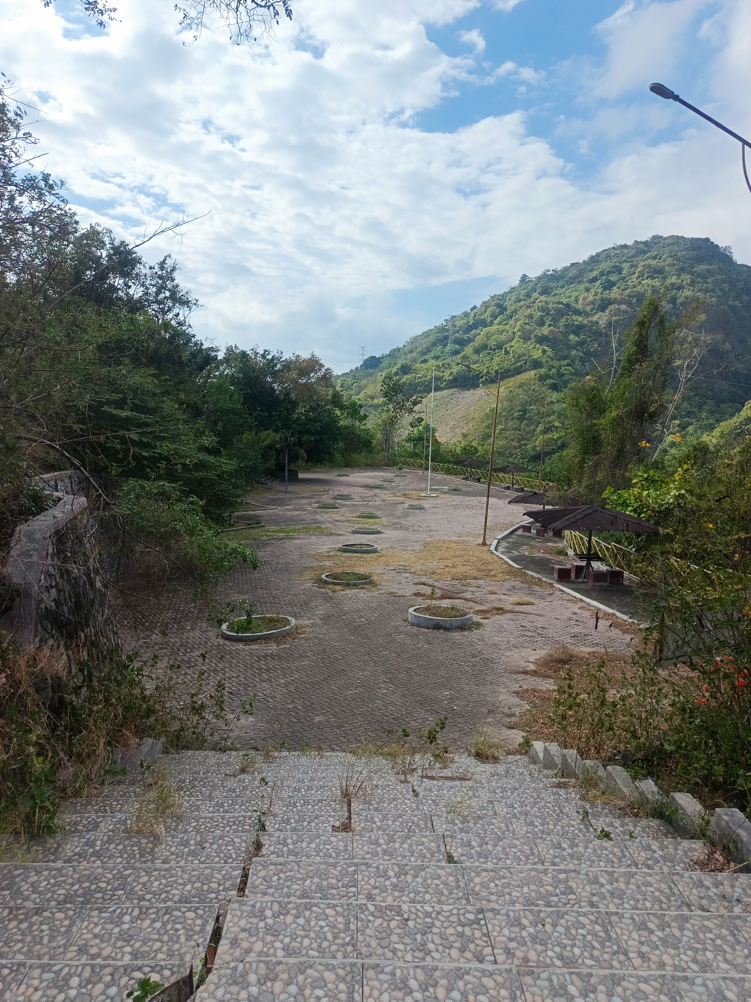 An outdoor, paved area with a slight decline, likely a recreational or parking space, is situated in a hilly, green environment. In the foreground, a wide set of stone steps descends towards the paved area. The steps are made of small, colorful pebbles embedded in concrete. Some of the steps show cracks, and sparse, dry grass grows in them.
The paved area itself is composed of interlocking rectangular stones, mostly covered in dry leaves and sparse vegetation. Several circular, low concrete planters are interspersed across the paving, some containing small, struggling plants. Further into the scene, a row of picnic shelters with dark thatched roofs and picnic tables can be seen along a curved edge of the paved area, demarcated by a yellow railing. The shelters appear somewhat weathered.
The background is dominated by a lush, green, tree-covered hillside rising steeply. The sky above is partly cloudy with white, puffy clouds against a bright blue backdrop. A modern street lamp with a dark, angled arm stands tall on the right side of the paved area. In the distance, on the hillside, a tall transmission tower is visible. The overall impression is of an open, somewhat neglected public space within a natural, verdant landscape, potentially located in Gorontalo, Indonesia, given the hilly terrain and vegetation. There are no people visible in the image.