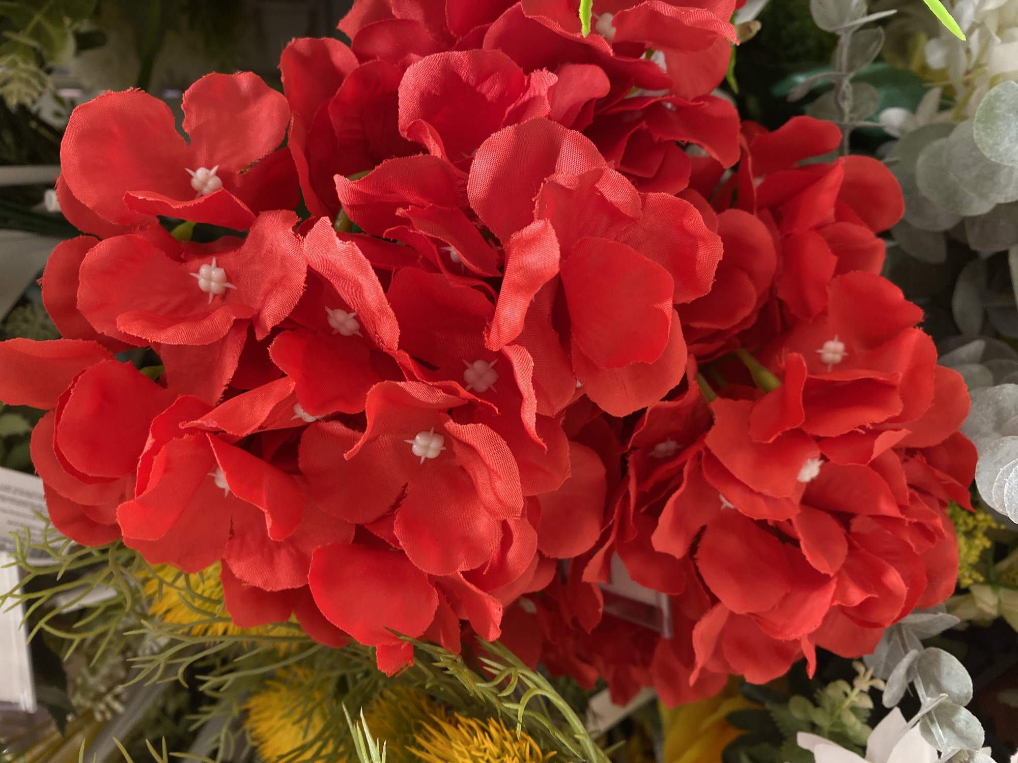 The image shows a close-up view of a bouquet of artificial red flowers. The flowers are clustered together, with some appearing in focus and others slightly blurred. Each flower has multiple petals, and in the center of some of the blooms, small white stamen-like structures are visible. In the background, there are hints of other artificial greenery and some yellow flowers. The lighting appears to be artificial, casting soft shadows on the petals. The setting is likely a retail environment, such as a florist shop or a home decor store, possibly in Đà Nẵng, Vietnam, given the request, although the image itself does not provide any direct clues to its geographical location. There are no people or animals present in the image. The focus is entirely on the texture and color of the artificial flowers.