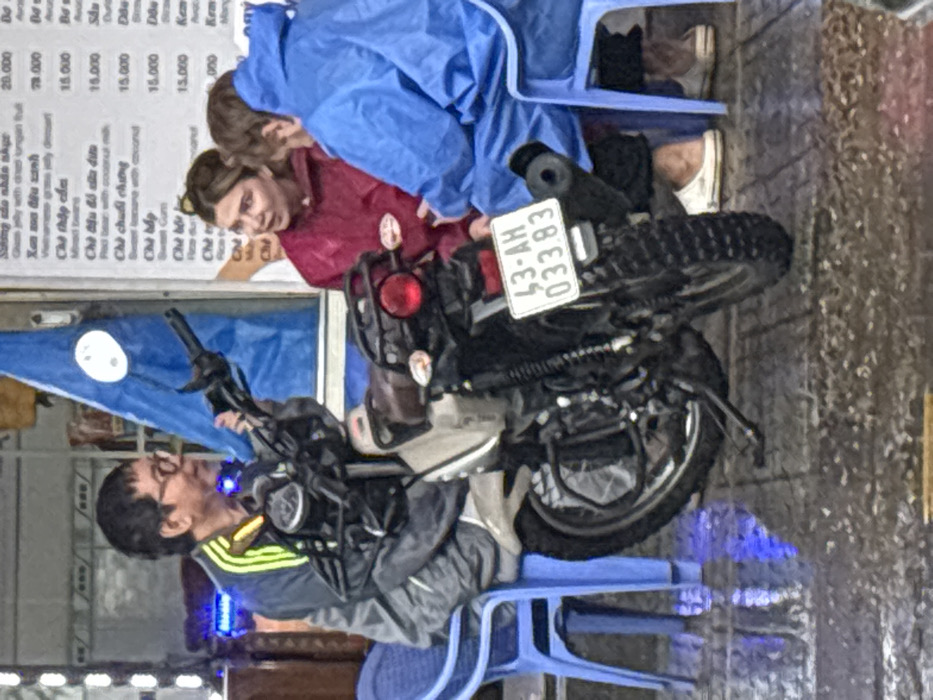 An outdoor scene in Ngũ Hành Sơn Ward, Đà Nẵng, Vietnam, depicts three individuals on a wet, tiled sidewalk. A grey and black motorcycle is centrally parked, displaying Vietnamese license plate '43-AH 033.83' on its rear fender. To the left of the motorcycle, an adult male, appearing of East Asian descent, wearing glasses and a grey tracksuit with yellow shoulder stripes, is seated on a blue plastic chair, looking towards the right. To the right of the motorcycle, a Caucasian female with light brown hair, wearing a maroon rain poncho, sits on a blue plastic chair, facing another individual in a blue rain suit, also seated on a blue plastic chair. All three appear engaged in conversation. A menu board with Vietnamese text and prices is visible in the background, behind the individuals on the right. The ground exhibits visible wetness and reflections, indicating recent precipitation. A blue flag or banner is partially visible in the upper background, behind the left individual.