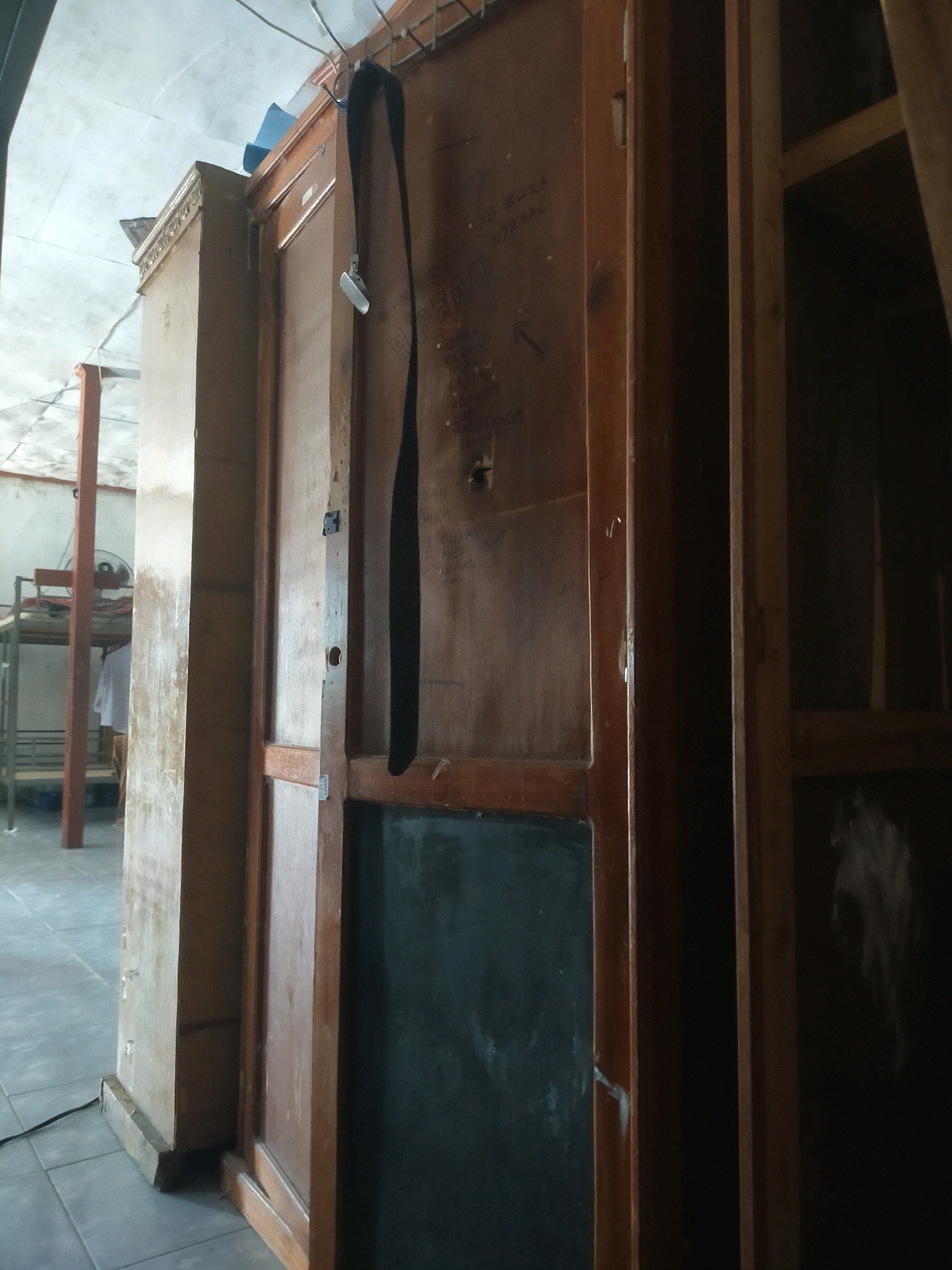 A dark wooden door with a black strap hanging from it is seen in a room. The door is part of a large wooden cabinet or closet. There is a black board panel on the lower part of the door. In the background, a metal bunk bed with a white shirt hanging from it is visible. The floor is tiled. The scene appears to be in Pekanbaru, Indonesia, likely in a dormitory or a similar living space.