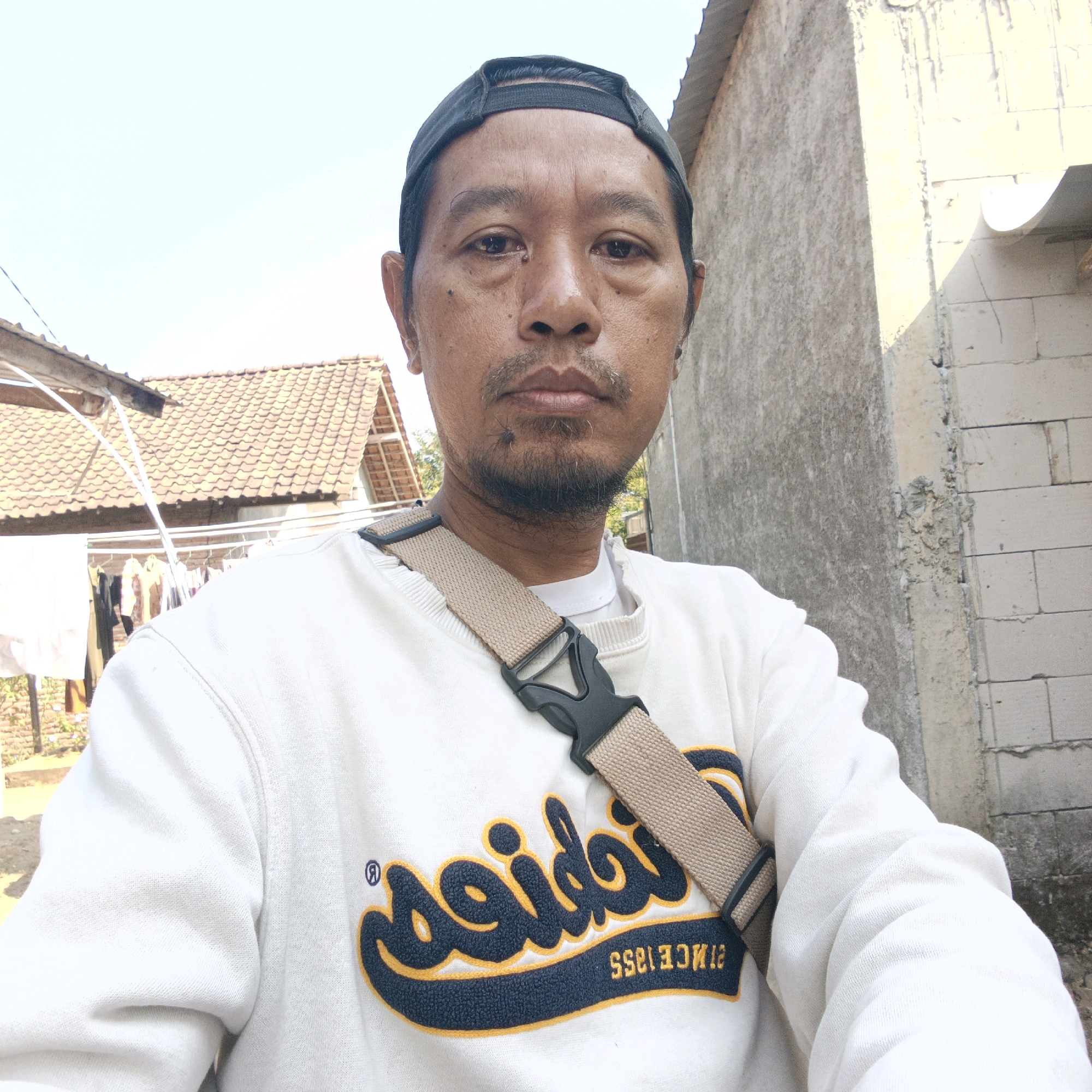 A close-up, eye-level, selfie-style shot shows a middle-aged Asian man from the chest up, outdoors in what appears to be a residential area in Tanahbaya, Indonesia. He is wearing a white crew-neck sweatshirt with a navy and yellow embroidered logo on the front, and a tan crossbody bag with a black buckle. He has short, dark hair, wears a black baseball cap backward, and has a mustache and goatee. The man's expression is neutral. Behind him, a blurred background shows residential buildings with red-tiled roofs, a clothesline with laundry, and a textured concrete wall. The lighting suggests it is daytime.