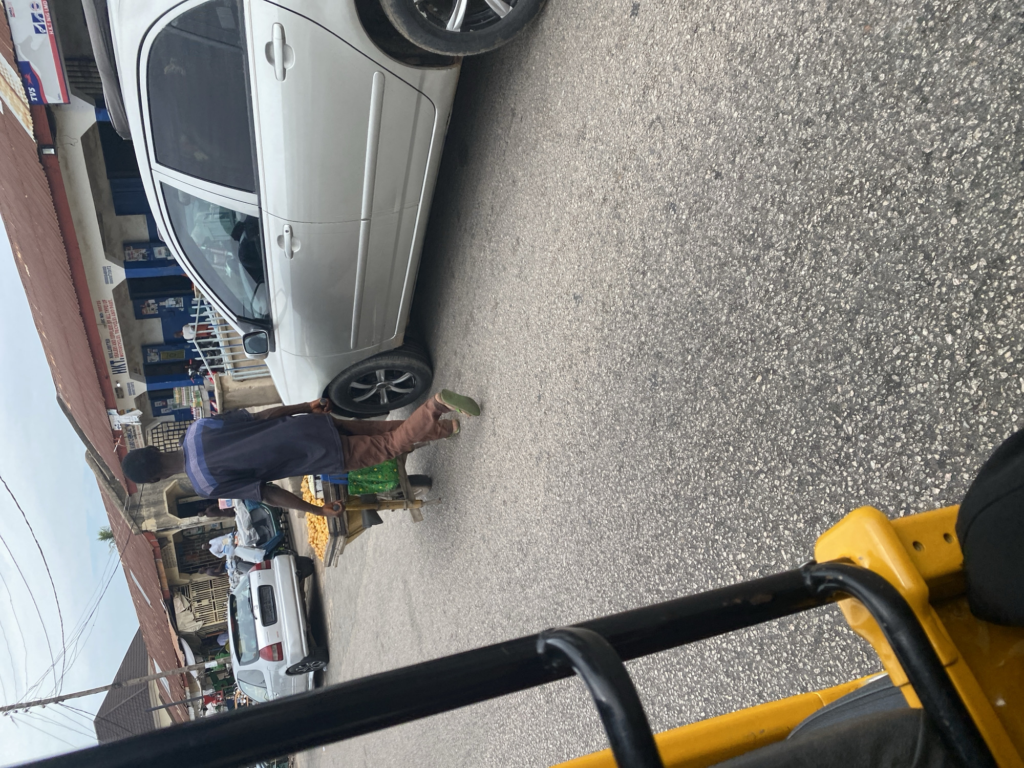A person is seen from behind, pushing a cart laden with goods, possibly produce, along a paved road. The cart has a yellow frame and a green tarp. The person is wearing a dark blue shirt and brown trousers, with light green shoes. They are walking alongside a white car. In the background, there are buildings that appear to be shops, with signage visible. One sign reads "TVS" and another, partially obscured, says "H.K.T NIG LIMITED DEALERS AND SUPPLIERS OF ALL TYPES OF MOTORCYCLES TRICYCLEI AND PART". The scene takes place outdoors on a paved road, suggesting an urban or semi-urban setting. The lighting indicates it is daytime. The overall impression is a street scene in a commercial area, likely a market or a road with shops.