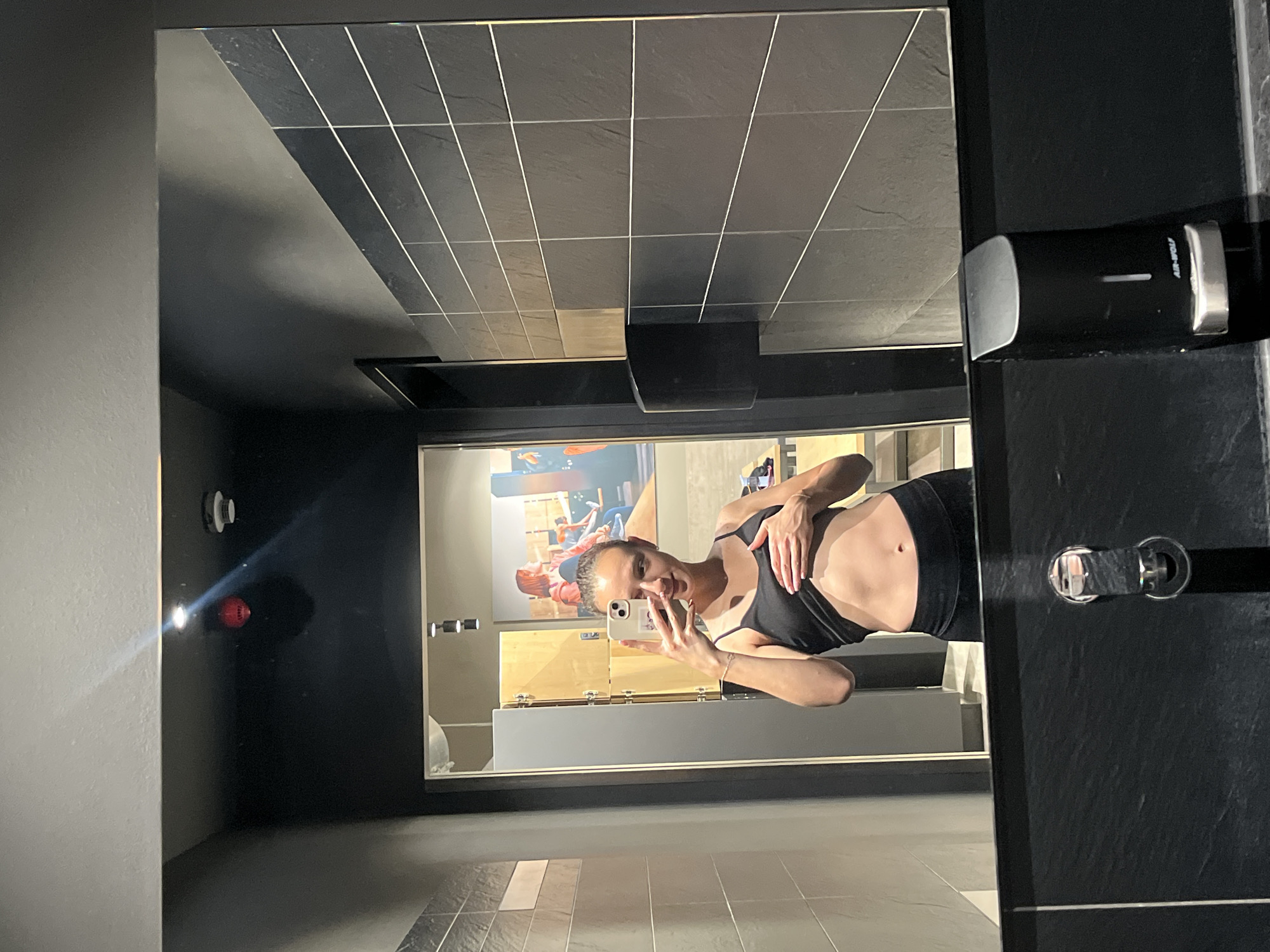A woman in workout attire takes a mirror selfie inside what appears to be a bathroom. She is wearing a black sports bra and black shorts. The bathroom features black tile floors and walls, with a sink and soap dispenser visible on the right side of the frame. The mirror reflects a scene of lockers and workout posters, suggesting a gym setting. While the image hints at an interior in Saarbrücken, Germany, no specific landmarks or items are present to confirm the exact location.