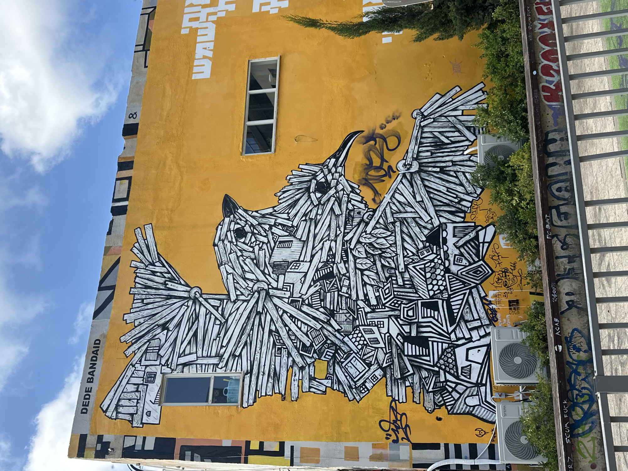 This is a medium shot, eye-level, exterior, daytime photograph of a building facade in Tel Aviv, Israel, featuring a large mural. The building is painted a vibrant orange and a section of the top is a light gray concrete with black and yellow geometric designs. The mural depicts two birds, intricately rendered in black and white lines, appearing to be constructed from wood planks and architectural elements like windows and rooflines. The bird on the left is larger and faces left, while the bird on the right is smaller, facing right and appearing to be pecking at something on the larger bird. Below the mural, two air conditioning units are visible, mounted on the wall, with dense green foliage obscuring the lower portion of the wall. Above the mural, on the orange section, white lettering in a stylized font is visible. On the gray concrete section, the words "DEDE BANDAID" are written in black capital letters. The background shows a bright blue sky with scattered white clouds. In the foreground, a metal railing with a wooden top is visible, with various graffiti tags painted on the wall behind it. The overall scene conveys an urban environment with a strong artistic presence.