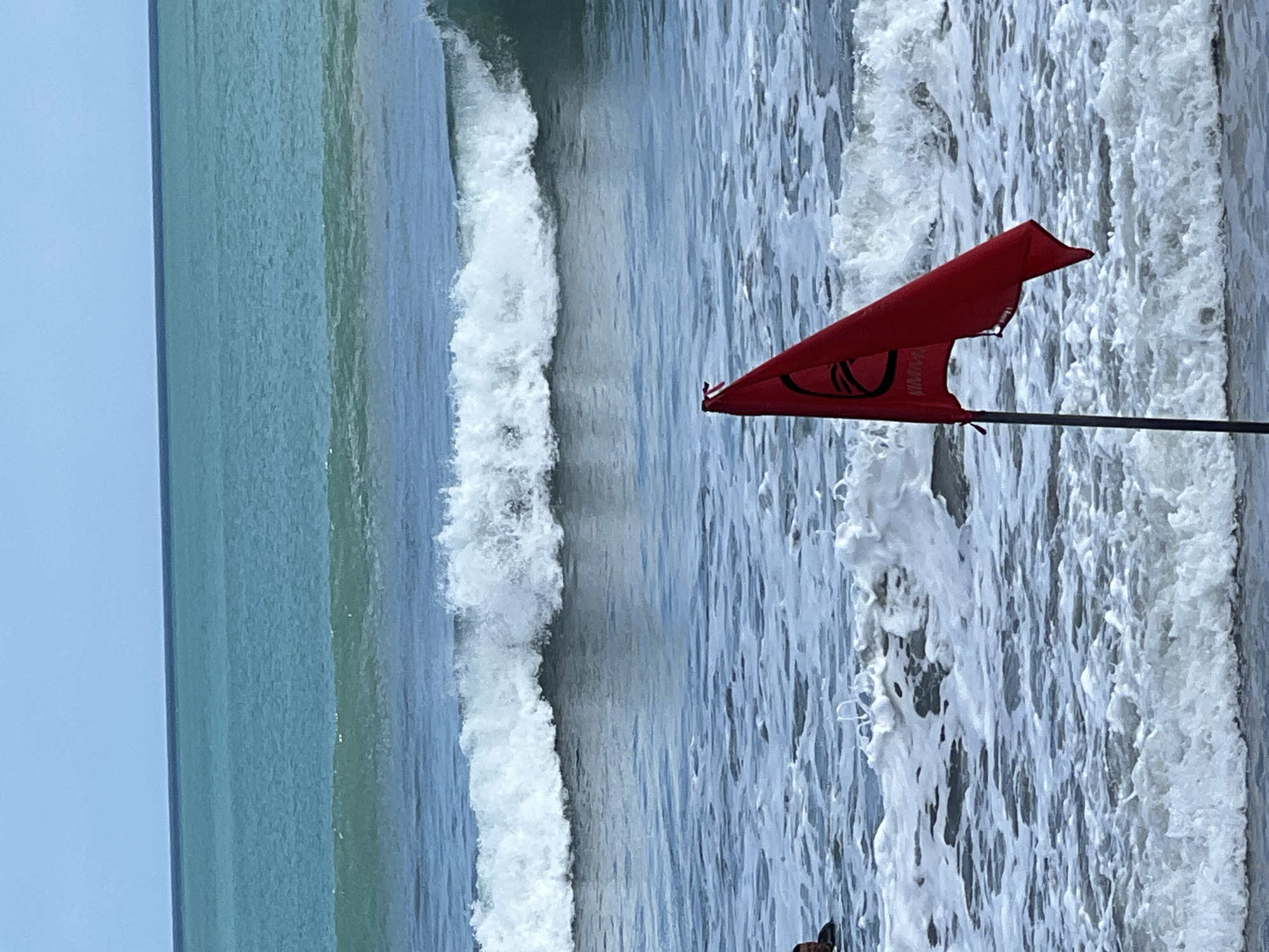 A red flag with a black circular emblem and text is planted in the choppy waters of Kuta Beach, Indonesia. The flag is on a dark pole and appears to be a warning of dangerous conditions. Waves are breaking in the foreground and midground, with a larger wave forming in the distance. The water is a deep blue and turquoise, with a clear blue sky above. A person is partially visible in the water on the left side of the image, possibly swimming or surfing. The scene depicts a dynamic ocean environment with a clear indication of potential hazards.
