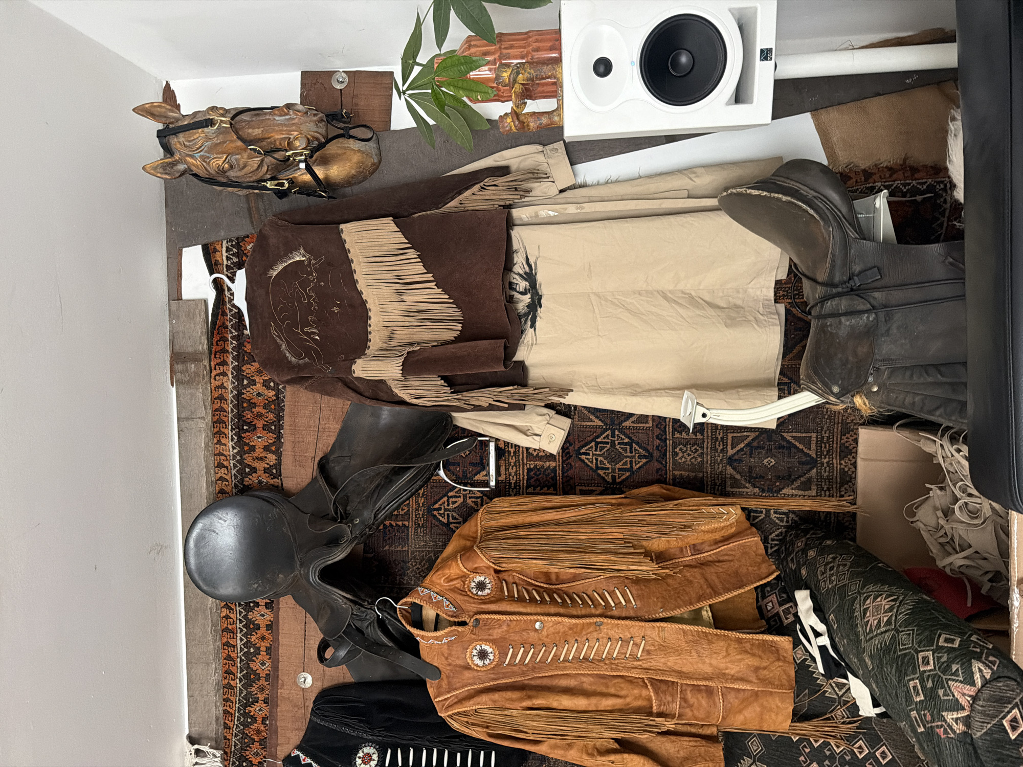 The image depicts a cluttered indoor space, possibly a storage area or workshop, with various equestrian and western-themed items. Prominently displayed are two saddles, one black leather saddle is hung high on the wall, and another darker brown leather saddle rests on a shelf lower down. Several garments are also visible, including a brown suede jacket with fringe and Native American-inspired embellishments, a tan-colored dress or skirt, and a black jacket with beaded details. A decorative wooden horse head with a bridle is mounted on the wall. Various rugs with geometric patterns are used as backdrops and to cover objects. A white speaker on a stand is positioned in the right side of the frame, next to a potted plant and a rustic vase. Boxes and rolled-up fabric are also present, contributing to the sense of disarray. The lighting suggests an indoor environment with artificial light. The scene is consistent with a collector's display or a storage space for vintage clothing and equestrian gear. There is no indication of the location in the city of Haarlem, Netherlands, or any people.