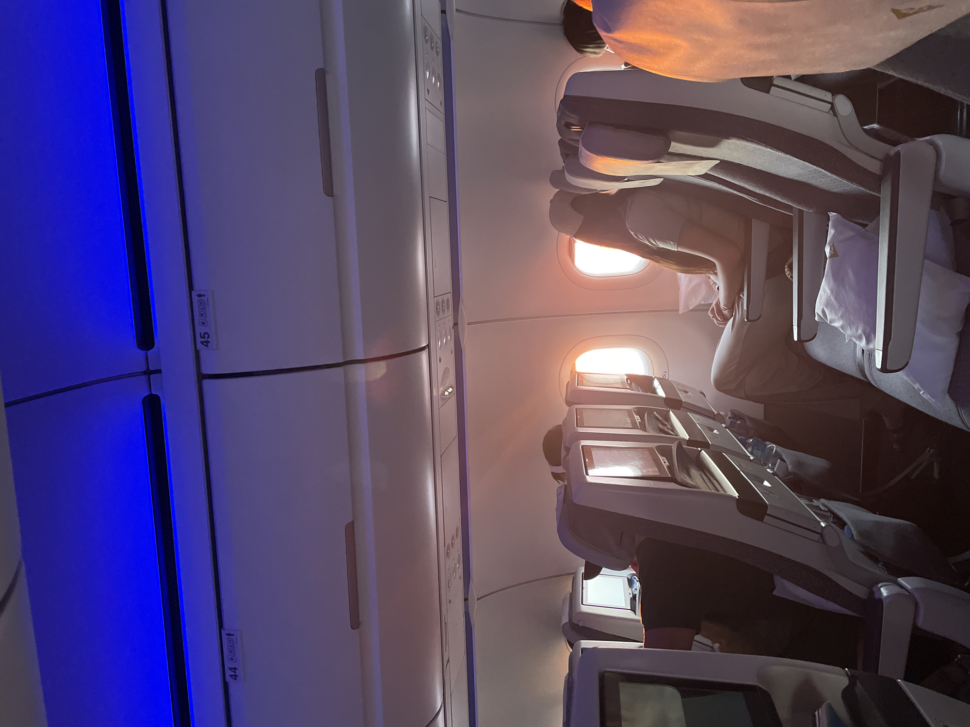 The image depicts the interior of an airplane cabin, likely during twilight or sunrise, given the warm orange glow illuminating the scene from the windows and reflecting off the surfaces. Several passengers are visible seated in their rows. In the foreground and mid-ground, seats with grey and white striped upholstery are prominent. Some seats have personal entertainment screens, which are either off or displaying a blank white screen. Overhead compartments are visible, with blue lighting emanating from above. Seat numbers '44' and '45' are visible on a compartment door in the upper left portion of the image. A person in the mid-ground is wearing a light-colored headscarf and a light-colored t-shirt, with their arms resting on their lap. Another passenger to their left is wearing a dark shirt. The overall environment suggests a typical airplane cabin setting. No specific details are present to indicate the location of Beykoz, Turkey.