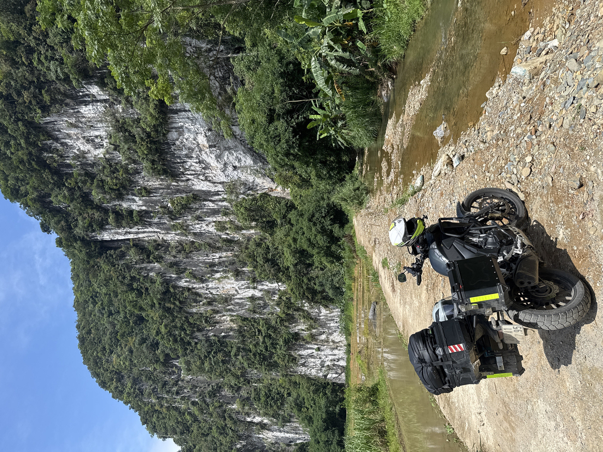 A motorcycle is parked on a dirt path next to a shallow, rocky stream. The motorcycle is outfitted with saddlebags and a top case, suggesting it is prepared for travel. A helmet rests on the motorcycle's seat. In the background, a sheer, forested cliff face rises dramatically, with patches of white rock visible. To the left of the path, a flooded rice paddy is visible, with tall green reeds growing along its edge. Banana trees and other lush vegetation flank the stream and the base of the cliff. The sky is clear and blue. The scene appears to be in a rural, natural setting, likely Pù Luông Commune, Vietnam, given its characteristic karst landscape and rice paddies. There are no people visible in the image.