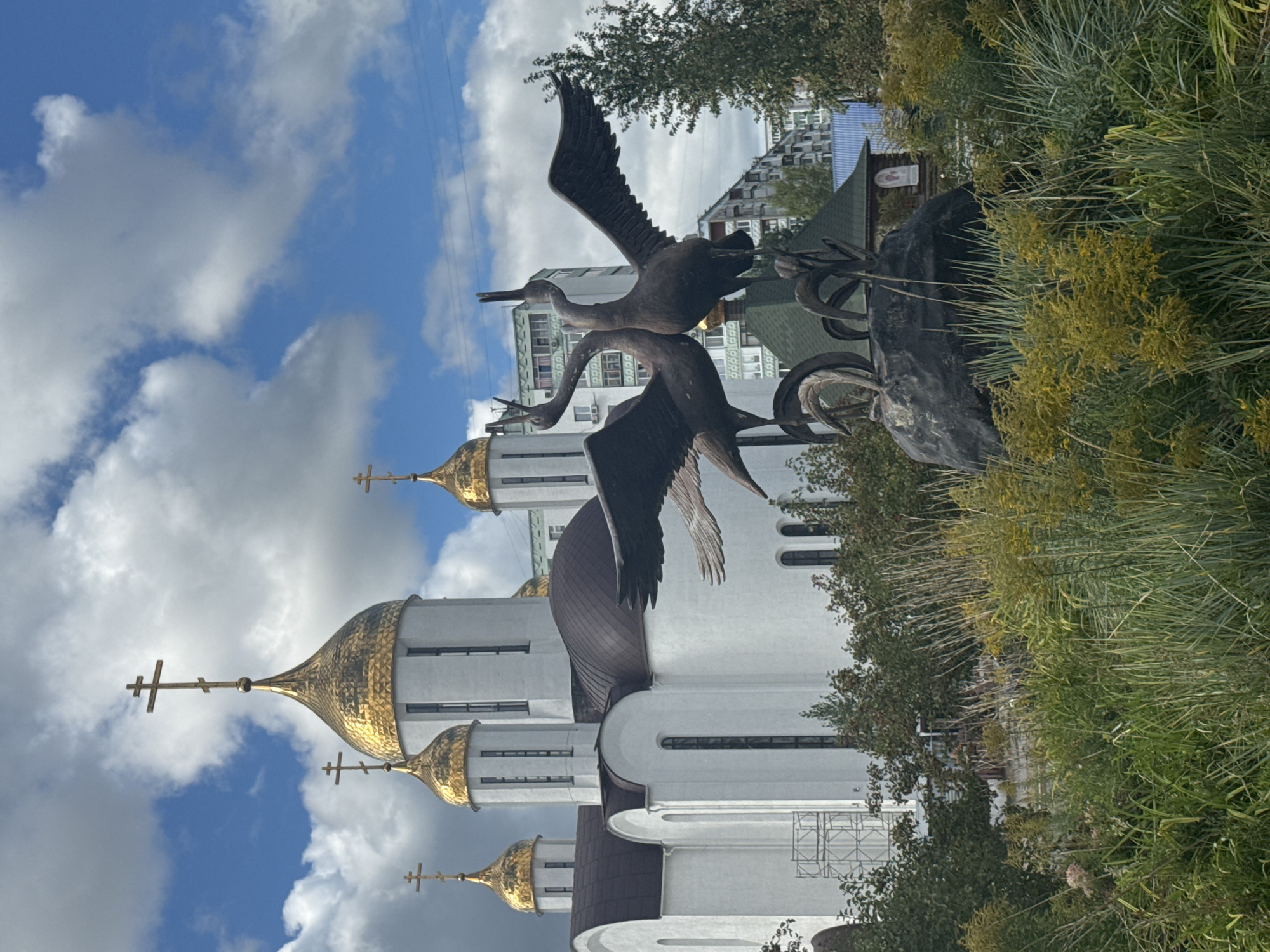 The image depicts a statue of two bronze cranes with wings spread, facing each other with their beaks almost touching, placed on a dark, rock-like base. The statue is surrounded by lush greenery, including tall grasses and smaller bushes, with yellow wildflowers visible. In the background, a white church with golden domes topped with crosses stands prominently, its architecture featuring arched windows and a large, dark-tiled dome. Adjacent to the church, several multi-story residential buildings are visible. The sky is a bright blue with scattered white clouds. This scene is located in Ufa, Russia, specifically at the Monument to the Cranes, near the Congress Hall Toratau.