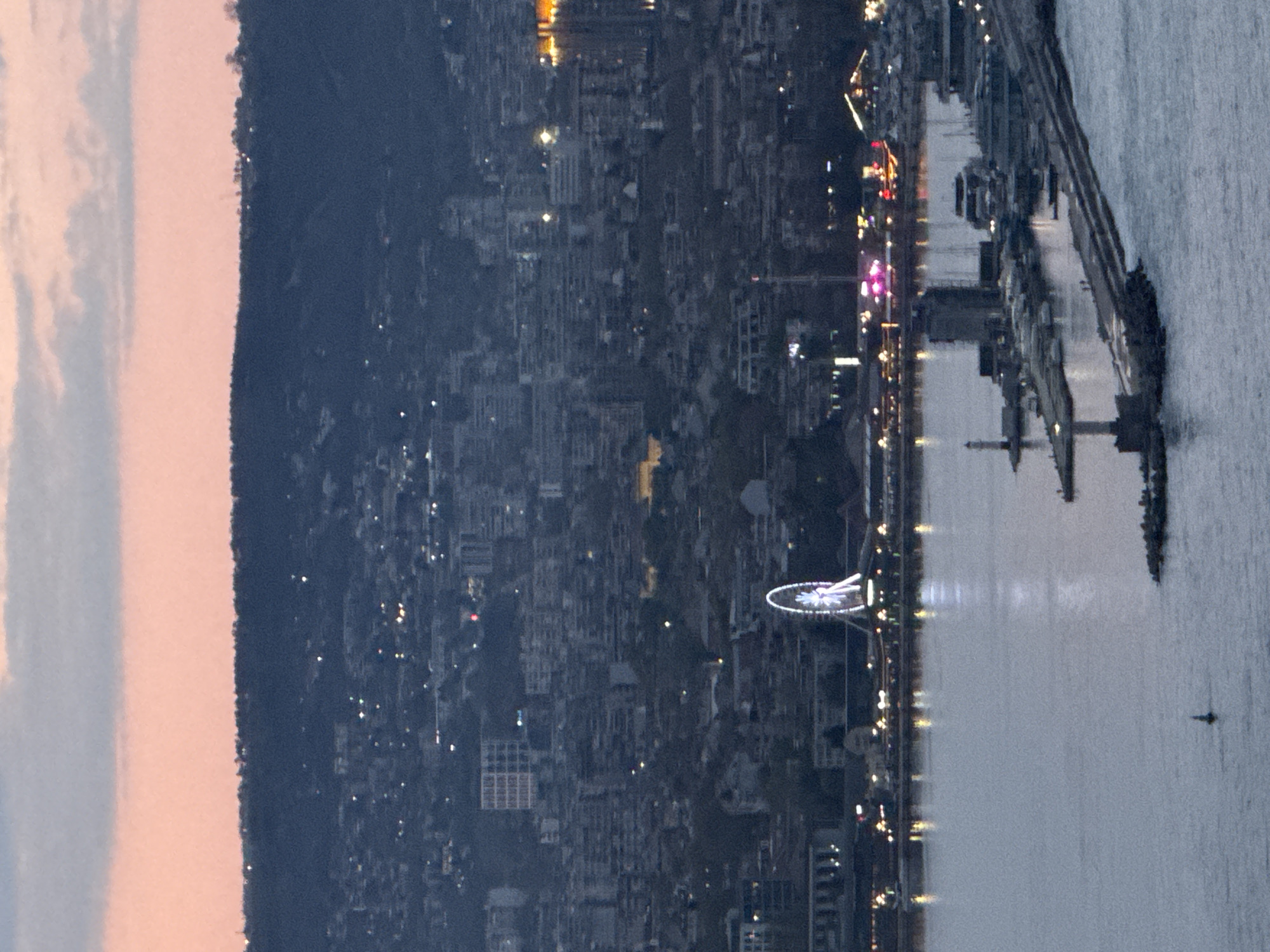 The image shows a cityscape at night in Varna, Bulgaria, viewed from an elevated position. A large, illuminated Ferris wheel is visible along the coastline. The city sprawls inland with many lights indicating buildings and streets. The sea reflects the lights of the city, and a pier extends into the water on the right side. A dark mountain range is seen on the left side of the city. The sky appears to have a hint of dawn or dusk coloring.