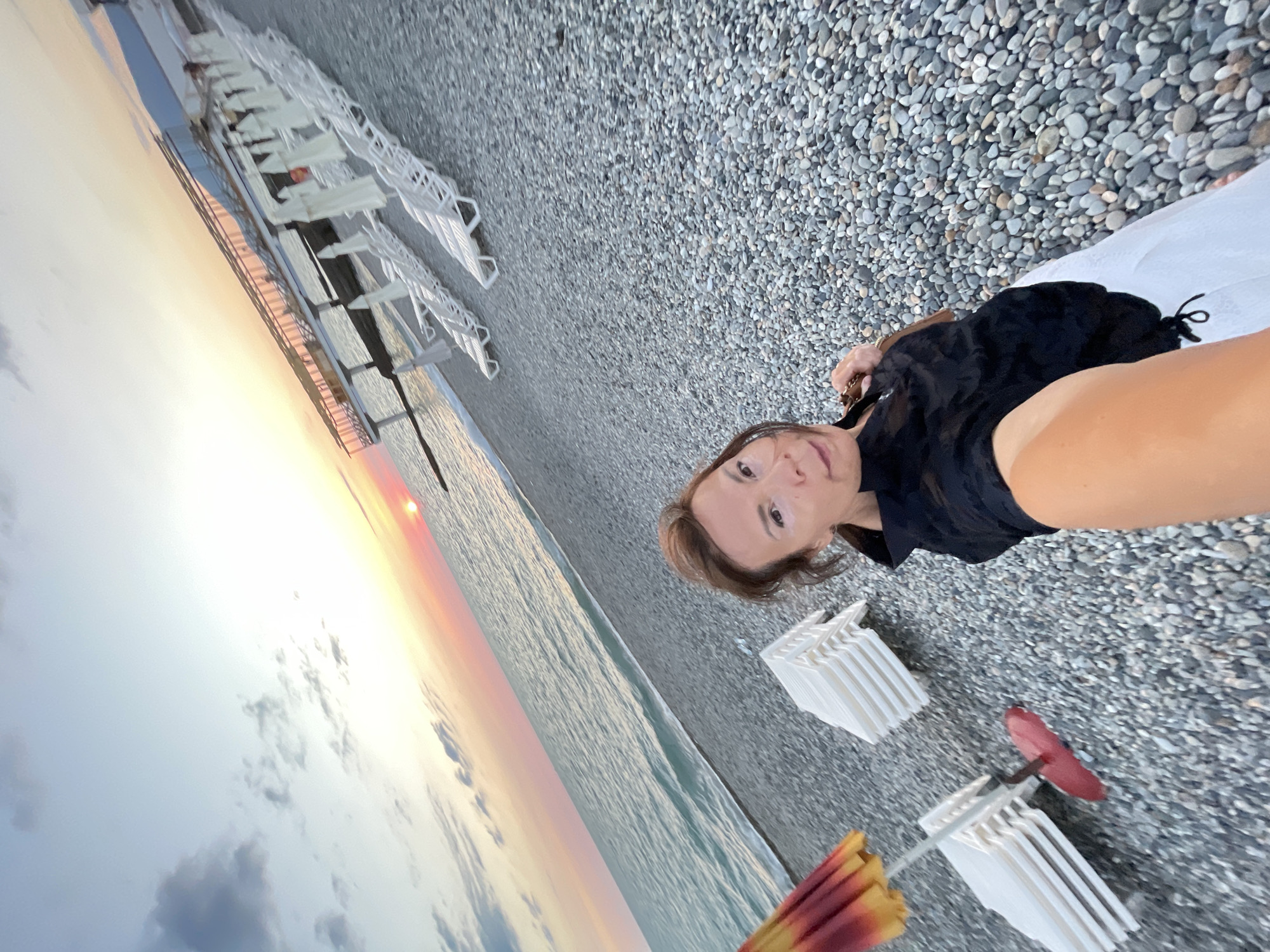 A woman stands on a pebble beach in Sochi, Russia, taking a selfie at sunset. The sun is low in the sky, casting a warm glow over the scene. The ocean is calm and a few clouds are visible. In the background, rows of empty lounge chairs and umbrellas are set up, suggesting a tourist area. The woman is wearing a black top and white skirt. The overall mood is peaceful and serene.
