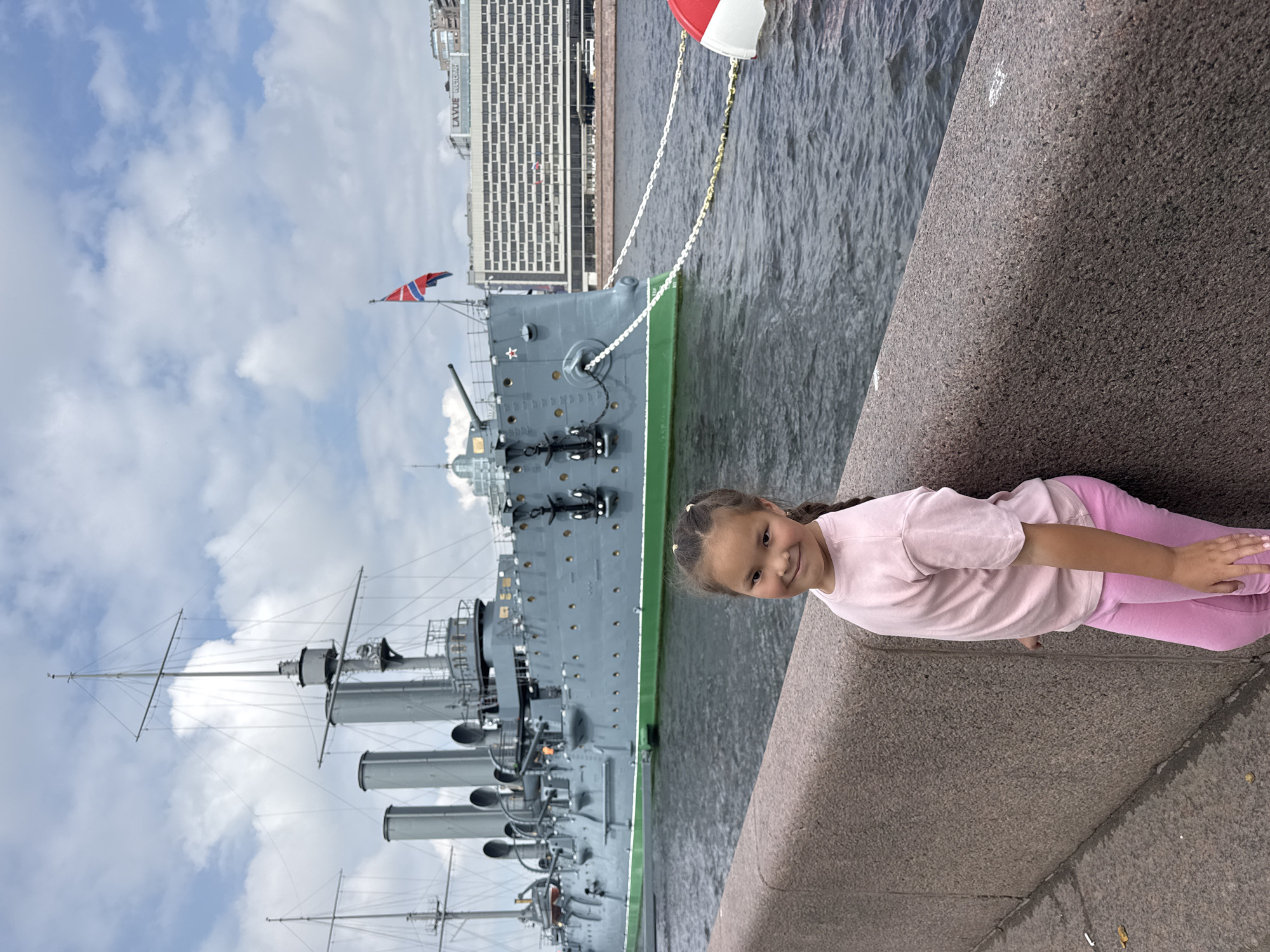 A young girl with her hair in a braid, wearing a pink t-shirt and pink leggings, smiles at the camera as she stands next to a granite embankment. Behind her, the armored cruiser Aurora, now a museum ship, is docked on the Neva River in Saint Petersburg, Russia. The ship, painted grey with a green waterline, has multiple smokestacks and masts. In the background, a modern hotel building with a sign reading "LAVUE PEKTOPAH" can be seen across the river. The sky is filled with clouds.
