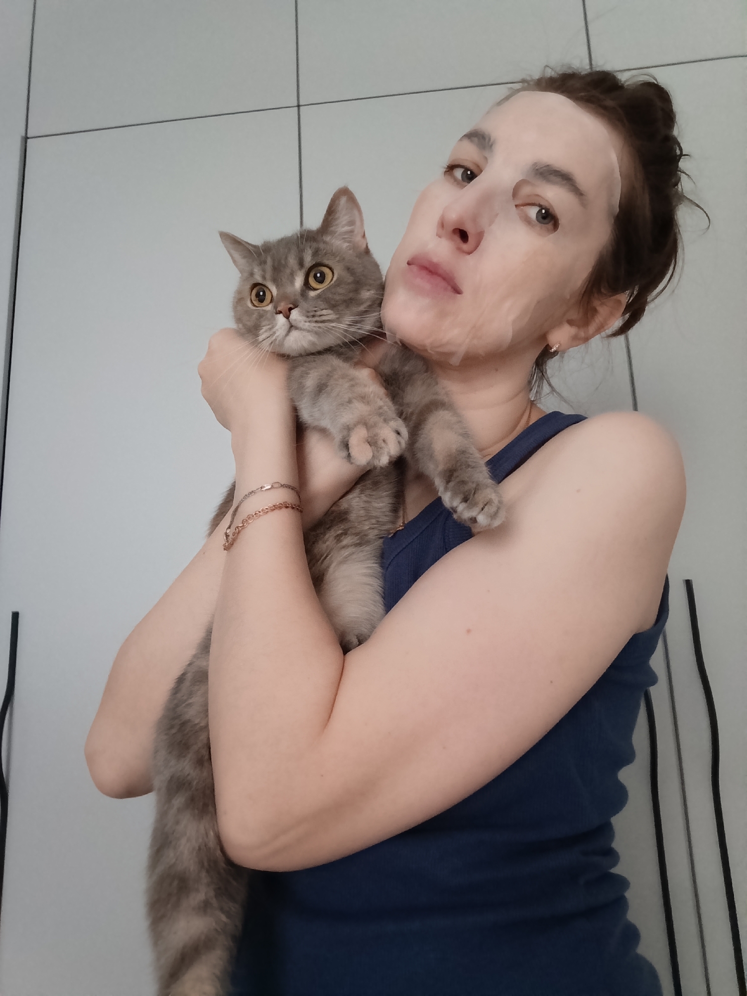 A woman in a blue tank top is holding a gray tabby cat. The woman has a white sheet mask on her face and her hair is pulled back in a bun. She is wearing two bracelets on her left wrist. The cat is looking to the left with its mouth slightly open. The background is a white wall with vertical black lines. The location is Chaikovsky, Russia.
