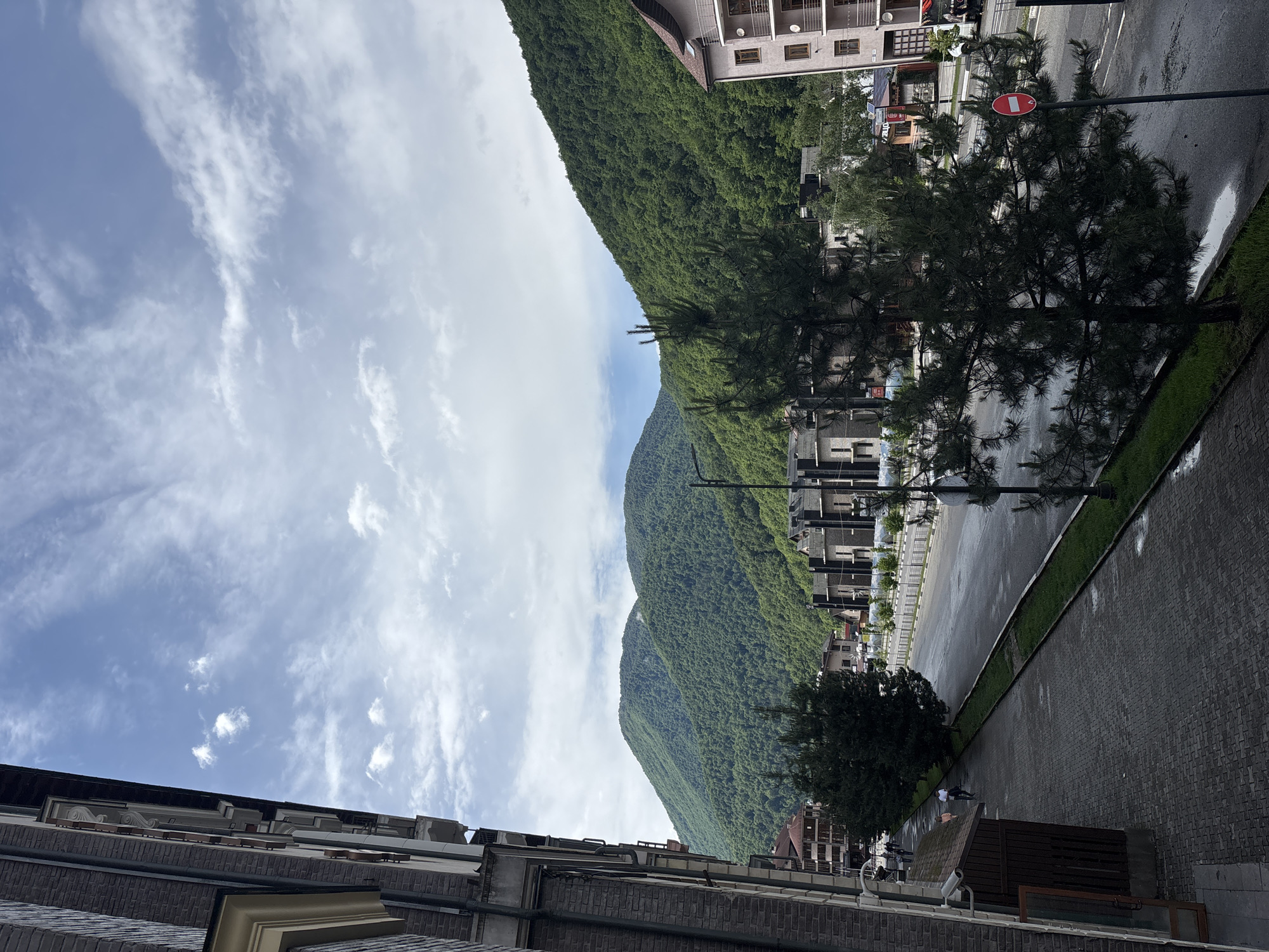 The image depicts a view of a street in Estosadok, Russia, with lush green mountains in the background. The foreground features a building on the left, a sidewalk with paving stones, and a street with wet asphalt. Several trees are scattered along the sidewalk and near the street. In the mid-ground, there are modern buildings with dark facades and balconies. The sky above is partly cloudy. A traffic sign, a red circle with a white horizontal bar, is visible on the right side of the street. A few people are walking on the sidewalk. The overall impression is a serene mountain town scene.