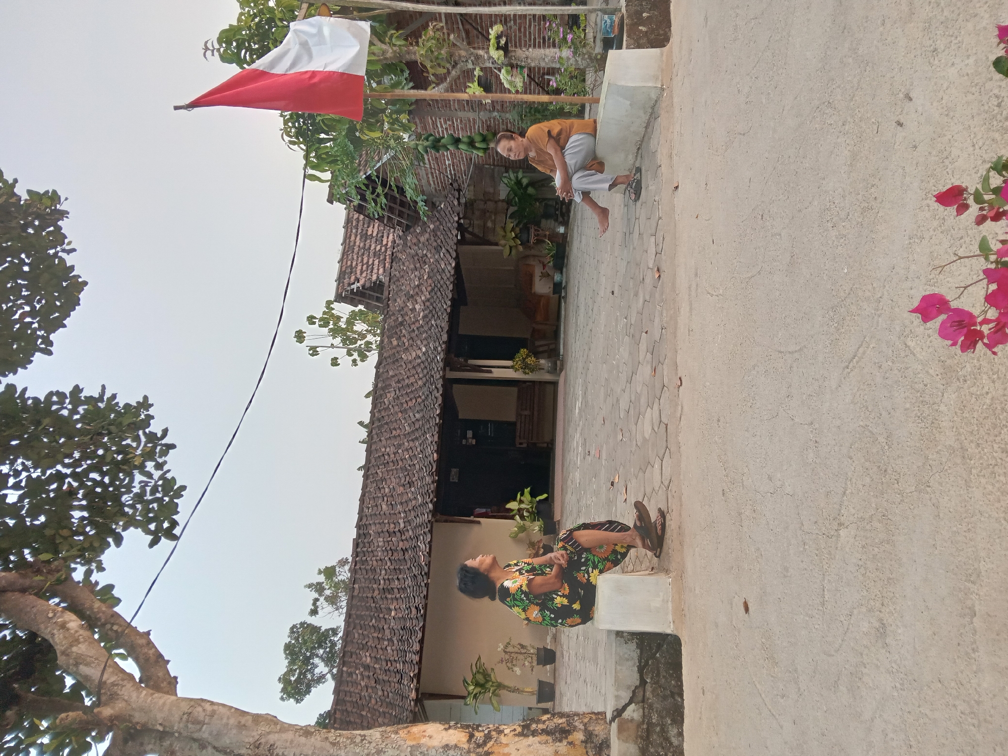 In the city of Kedunggudel, Indonesia, two elderly women are seated on concrete blocks outside a traditional Indonesian house. The woman on the left wears a floral dress and is looking to the right. The woman on the right is wearing a brown shirt and light-colored pants, and is looking forward. The house behind them has a red tiled roof and appears to be made of brick and wood. A red and white Indonesian flag is visible on a pole to the right of the house. Trees and potted plants surround the area, and the ground is paved with hexagonal stones. The sky is clear and pale, suggesting it is either early morning or late afternoon.