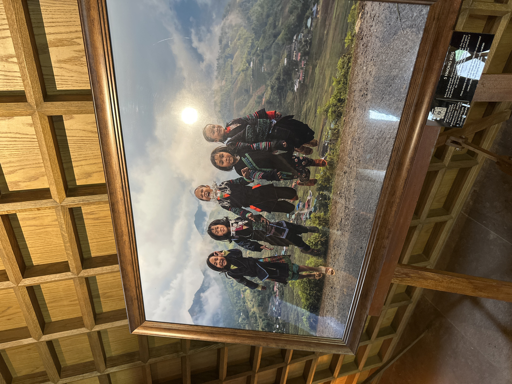 A framed photograph, displayed on a dark wooden easel, depicts five young children from the Hmong ethnic group joyful and mid-air, seemingly jumping on a dirt path overlooking a mountainous landscape in Púng Luông, Vietnam. The children, dressed in traditional dark garments with vibrant embroidered patterns, exhibit wide smiles and open-mouthed laughter, positioned against a backdrop of green terraced hills, a scattered village, and mountains receding into a cloudy sky. A bright sun or light reflection is prominent in the upper right portion of the photograph. The wooden frame of the artwork features a decorative profile, showing minor surface scratches.
The easel stands on a dark tiled floor, in front of a wall clad with light brown wooden panels arranged in a grid of recessed squares. A black informational plaque is affixed to the easel's lower support bar, identifying the image as "5 HMONG KIDS" and providing a brief description of the scene, stating the children are "jumping on a road that needed no directions to experience their childhood." The plaque also details print availability, limited edition information, and contact details for "PRECIOUS HERITAGE ART GALLERY SAIGON" in HCMC, including a QR code and website. Reflections from an unseen light source are visible on the surface of the framed photograph.