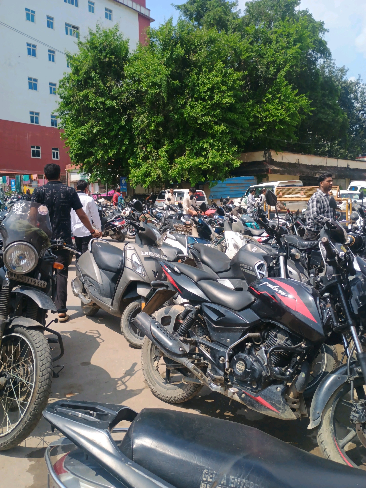In Varanasi, India, a sunny day unfolds in a crowded parking area filled with motorcycles and scooters. A large, multi-story building with white and red accents forms the backdrop, its numerous windows reflecting the light. Lush green trees partially obscure the building, adding a touch of nature to the urban scene. In the foreground, a dark motorcycle with a reflective windshield stands prominently, its license plate reading "KA-05KY-888". To its right, a grey scooter, its license plate bearing "UP-65DY-5340", is parked. A black motorcycle with red accents, possibly a Bajaj Pulsar, is also visible. The ground is a mix of asphalt and dirt, marked with shadows cast by the vehicles. People, dressed casually in shirts and pants, are dispersed among the parked vehicles, some walking and others attending to their motorcycles. The atmosphere appears busy and functional, typical of a public parking space in a bustling Indian city.