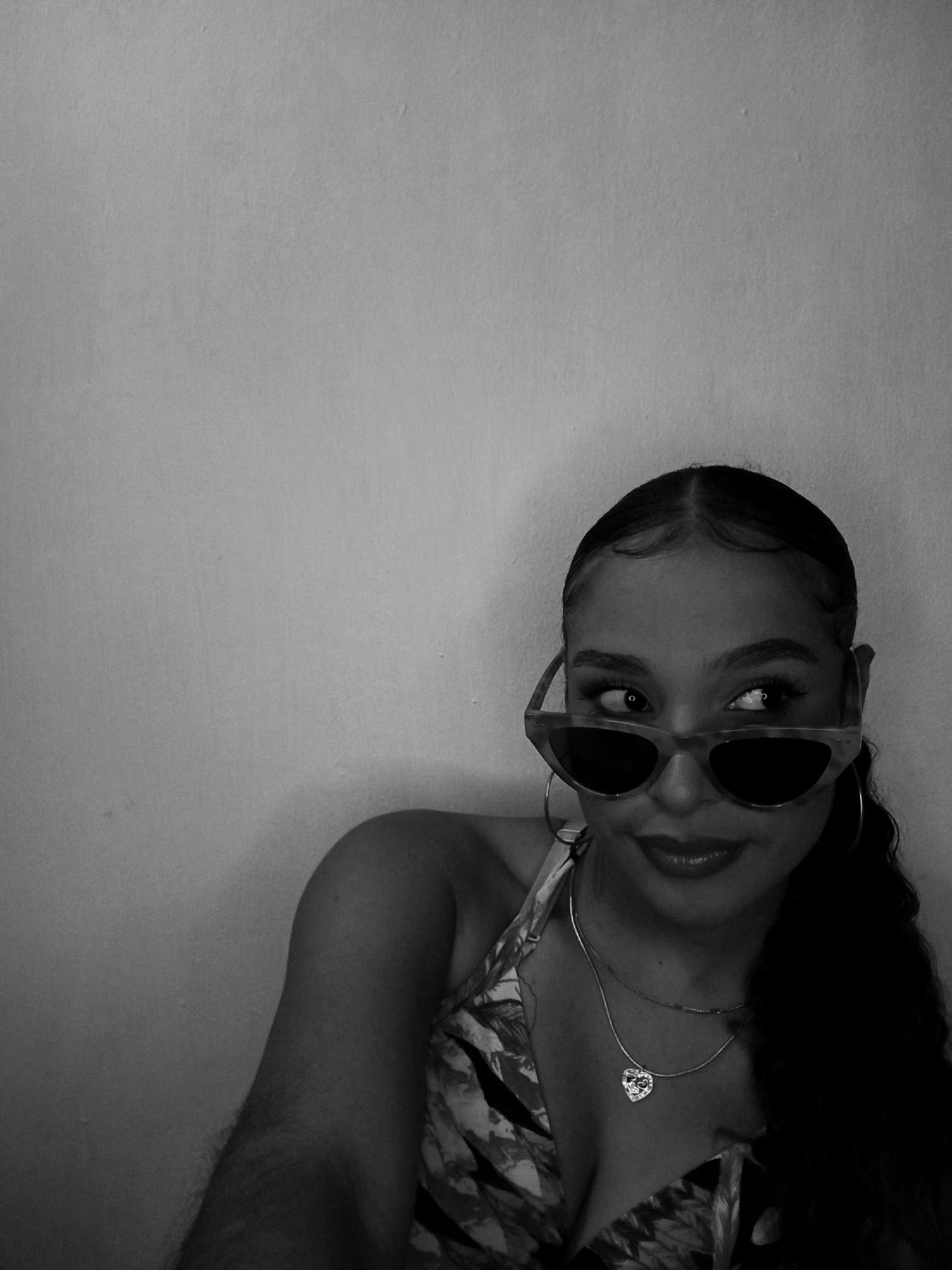 The image is a black and white portrait of a woman wearing sunglasses and jewelry. She is looking to her left. The background is a plain wall. The location is not discernible from the image and no information is provided to suggest the photo was taken in Juan Griego, Venezuela.