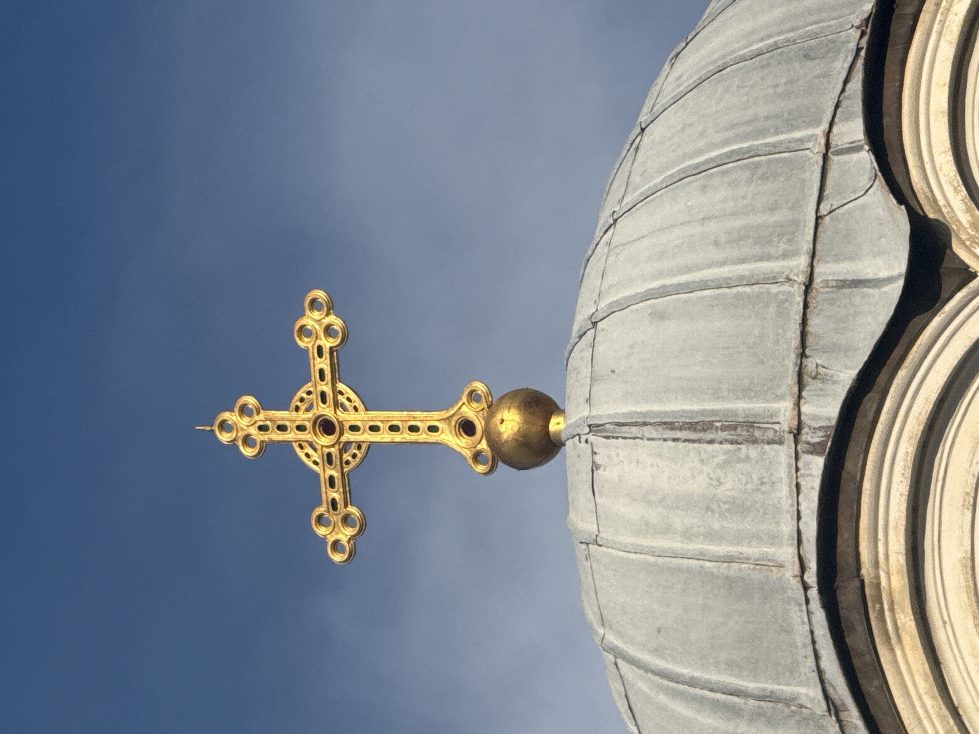 The image displays a close-up, upward-looking view of an ornate golden cross affixed to the apex of a dome against a clear blue sky. The original image is rotated 90 degrees, showing the cross pointing to the left and the dome on the right.
When viewed in its intended upright orientation, a meticulously crafted golden cross stands vertically. It features intricate detailing, including circular embellishments at the ends of its arms and decorative patterns along its length. Small, dark red or purple-hued gems are embedded in the cross, notably at its center. A polished golden sphere connects the base of the cross to the dome structure.
The dome itself is grey and appears to be constructed from segmented panels, possibly metal, exhibiting distinct seams and a weathered texture. On the far right edge of the image, a portion of a lighter, multi-tiered architectural element is visible, suggesting a decorative collar or an adjacent section of the building. The background is a uniform, bright blue sky, indicative of clear weather during daytime. There are no people visible, nor any discernible text. The architecture points towards a religious edifice, consistent with the location context of Sofia, Bulgaria, likely an Eastern Orthodox church.