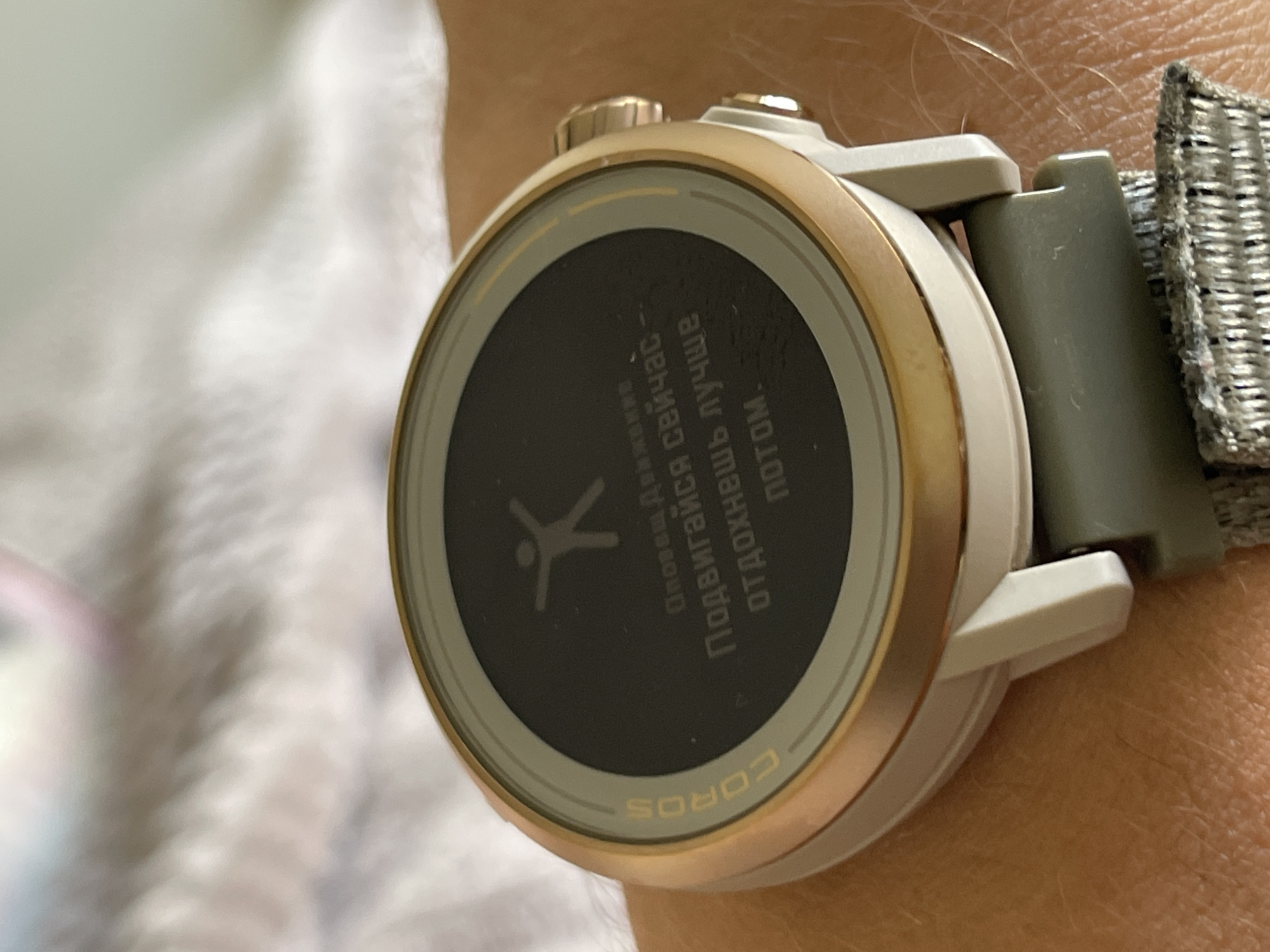 A close-up shot shows a COROS brand smartwatch worn on a person's wrist. The watch face displays a stick figure of a person, with text in Russian above and below the figure. The text reads "Подвигайся сейчас, Отдохнешь лучше потом," which translates to "Move now, You'll rest better later." The watch has a gold-colored bezel, light-colored case, and a woven nylon strap. The person's skin tone is light. The watch is the main subject of the image. The environment appears to be indoors.