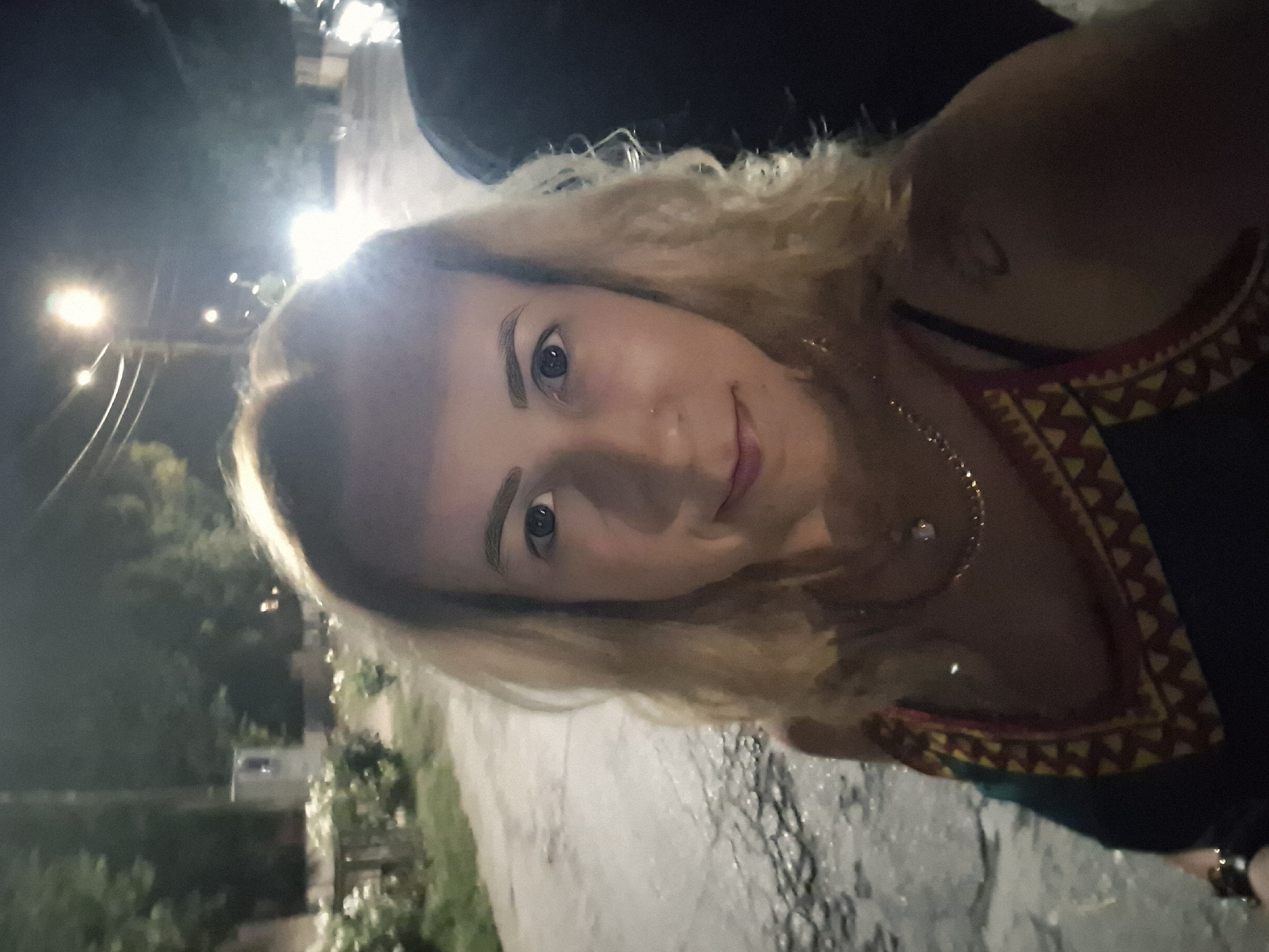 A woman with blonde, wavy hair and blue eyes is pictured in a selfie taken at night. She is wearing a dark-colored dress with a colorful patterned neckline and a thin necklace. A small nose ring is visible. The background shows a dimly lit street with some trees and streetlights. A portion of another person, dressed in dark clothing, is visible on the right side of the frame. The ground appears to be paved and somewhat uneven. The overall lighting suggests an outdoor urban environment at night.