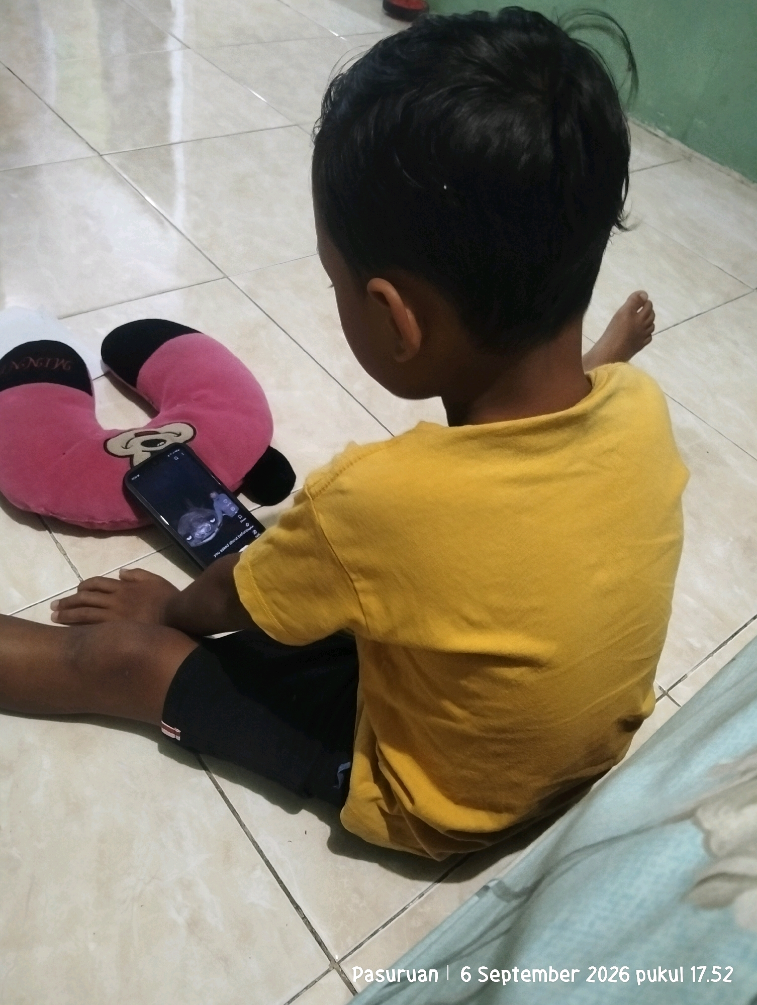 A young boy sits on a tiled floor, facing away from the camera, engrossed in a smartphone. He is wearing a yellow t-shirt and dark shorts. To his left, a pink U-shaped pillow with black accents rests on the floor. The phone displays an image of a cartoon character with text that reads "WHO ASKED ABOUT BEFORE." The environment appears to be indoors, with a green wall visible in the background. The bottom right corner of the image contains a timestamp and location: "Pasuruan | 6 September 2026 pukul 17.52".