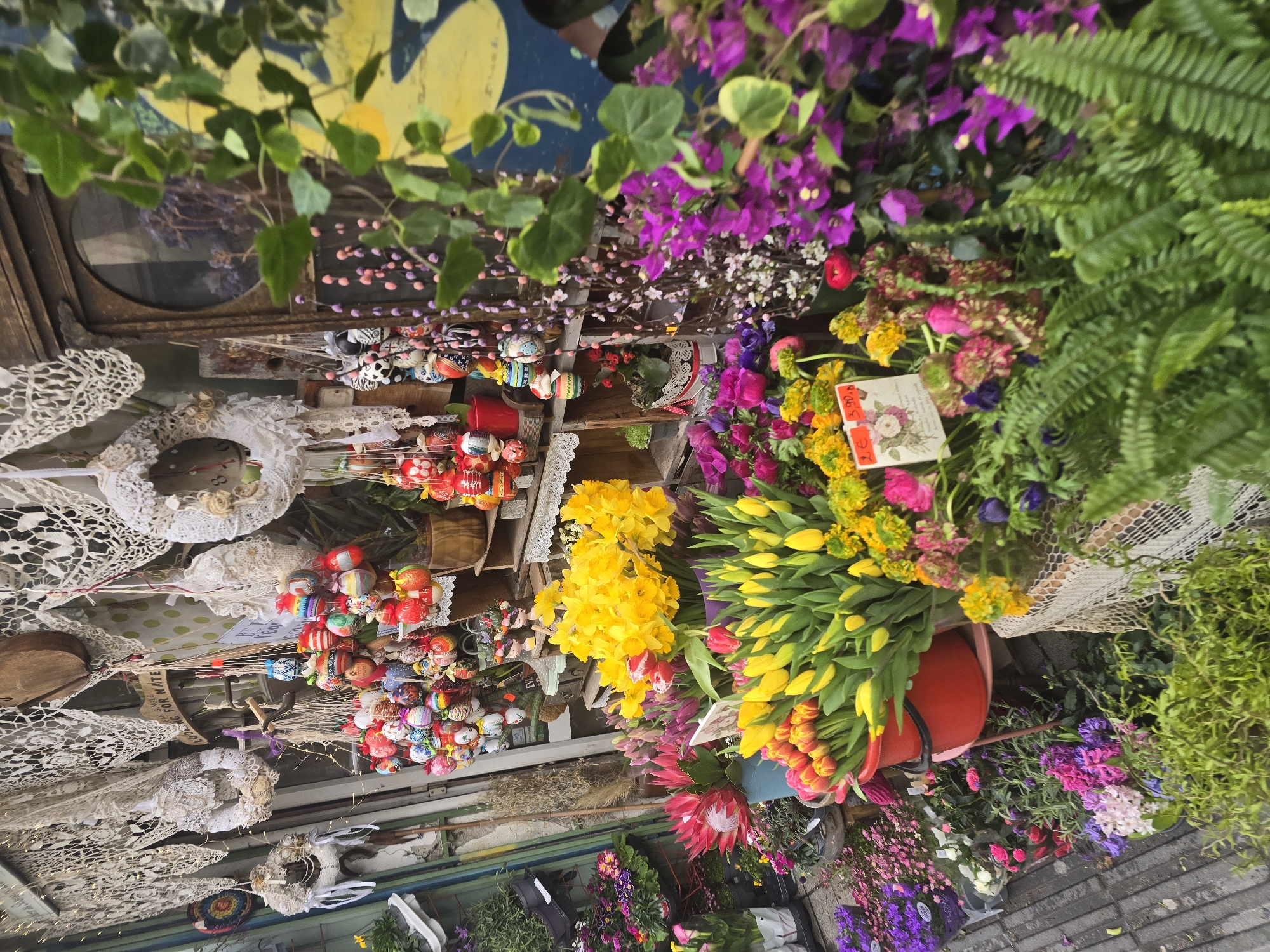 This is an eye-level view of the interior and exterior of a flower and gift shop. The shop appears to be in Sofia, Bulgaria, given the Cyrillic script on some price tags. The exterior and ceiling is decorated with an array of colourful flowers, hand-painted eggs, and doilies. There are buckets of tulips, daffodils, and bougainvillea. The shop sells a variety of handcrafted items, including lace doilies, floral arrangements, and decorated eggs. Some of the items are displayed on shelves, while others are hanging from the ceiling. The overall impression is of a quaint and charming shop that is full of character.