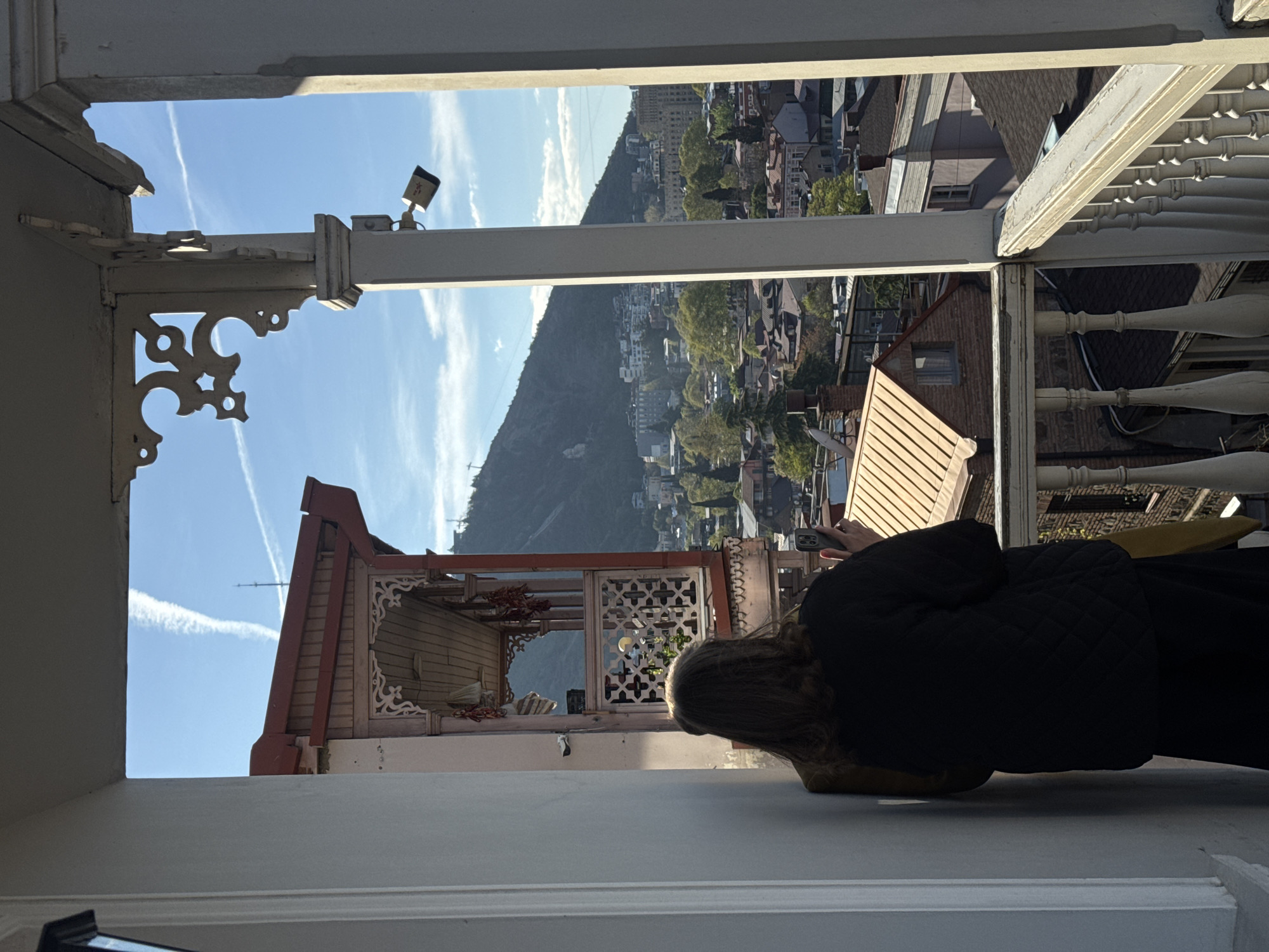 A woman is seen lying on a white surface, possibly a balcony or ledge, looking out at the city of Tbilisi, Georgia, through an ornate white wooden frame. The city view includes a large, tree-covered mountain, buildings with various roof colors, and a blue sky with contrails. A decorative, possibly historical, building with wooden details is partially visible to the left of the woman. The woman is wearing a black jacket and dark pants. She appears to be using a phone. The frame through which the view is presented is elaborately decorated with white carved details. A surveillance camera is mounted on one part of the frame.
