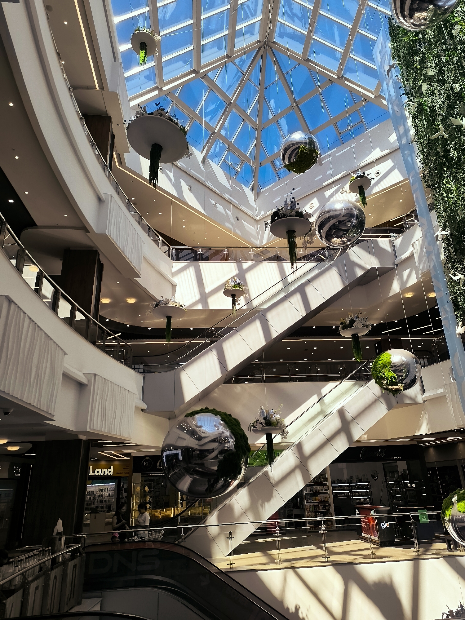 The image shows the interior of a multi-level shopping mall, likely illuminated by natural light filtering through a large glass ceiling. Several escalators are visible, connecting different floors. Suspended from the ceiling are decorative spherical objects, some of which appear to be filled with greenery. There are various shops and storefronts visible on the different levels, with some signage indicating "Land" and "glo". A few individuals are present in the mall, including two people standing near a shop and another person on an escalator. The architecture of the mall is modern, with clean lines and white surfaces. The overall impression is that of a bright and contemporary commercial space. There is no indication of the location being in Qarağandı city administration, Kazakhstan.