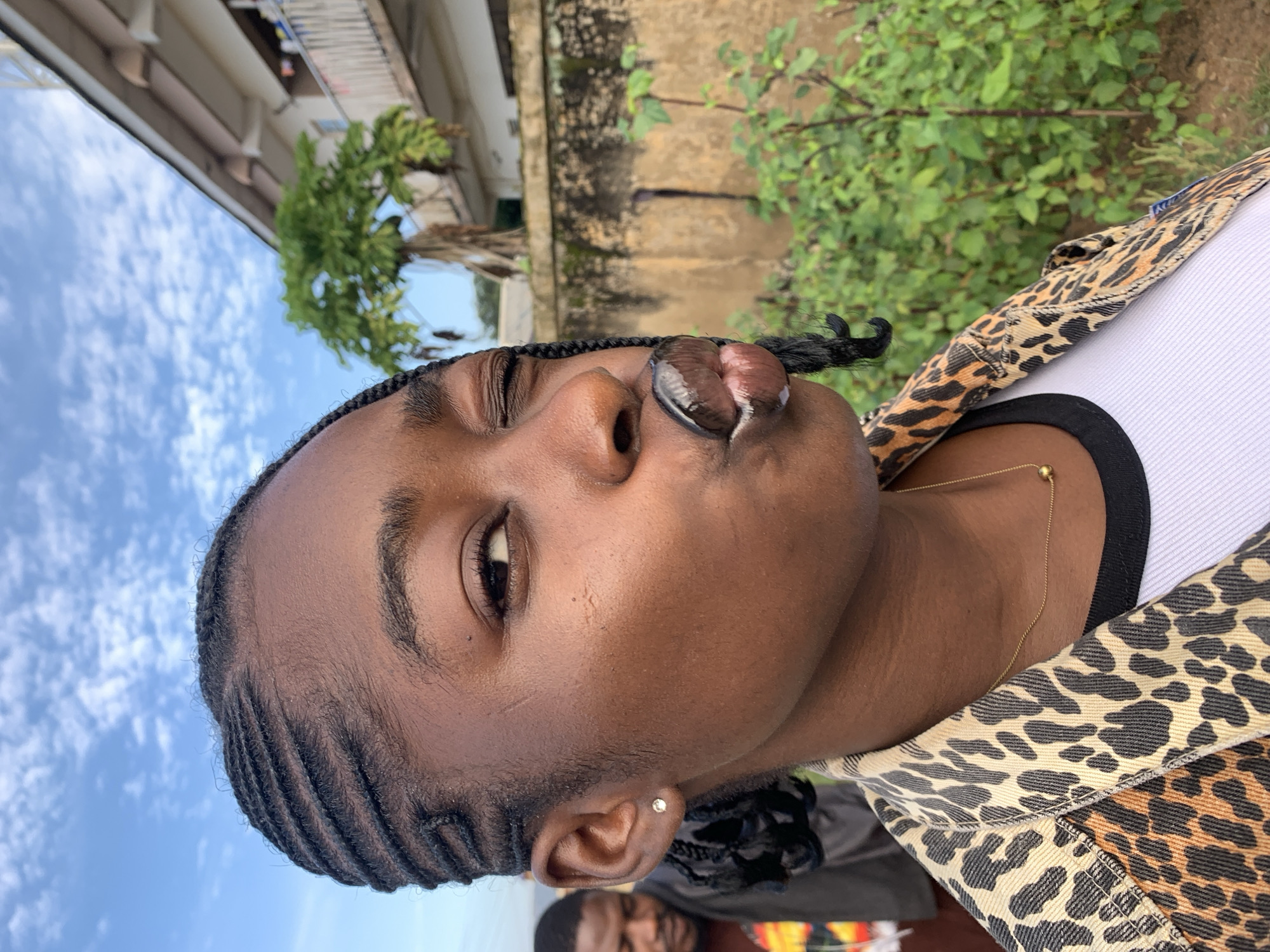 A young Black woman with braided hair and a leopard print jacket is captured in a close-up, outdoor selfie. She is winking, with her lips pursed as if blowing a kiss. A thin gold necklace with a small ball pendant is visible. In the blurred background, another person stands, and a building with a balcony and lush green vegetation are present. The sky is blue with scattered clouds. The image appears to have been taken in Rafin Par, Nigeria.
