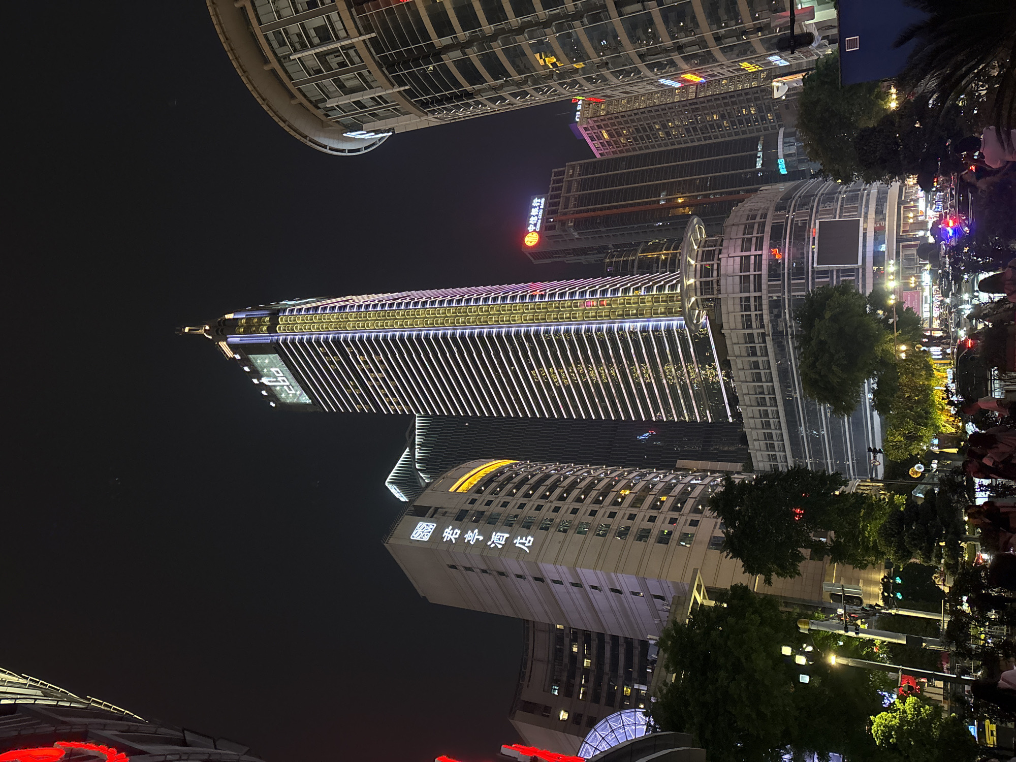 At night, a street-level view looks up at illuminated buildings in the Yuzhong District, China. Buildings of varying heights and shapes line the frame. The tallest building has thin vertical white lights spanning its height. A building with Chinese characters sits lower and to the left of the tall building. The right side of the view shows trees and traffic lights along the street. A crowd of people can be seen in the extreme right corner.