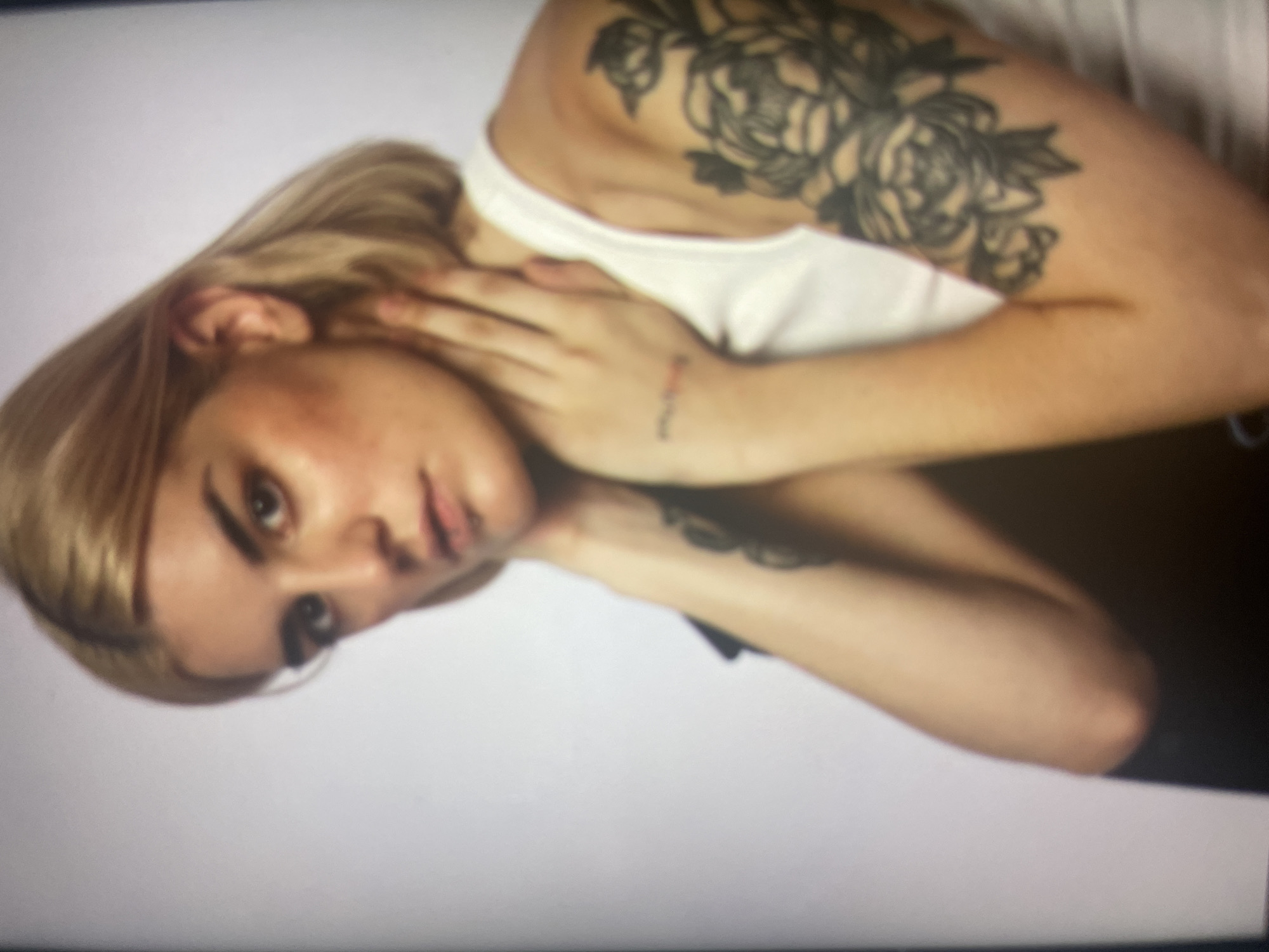 A medium shot portrait depicts a fair-skinned person with blonde hair and dark eyebrows, looking towards the viewer with a neutral expression. They are wearing a white tank top and have several tattoos visible on their arms. One prominent tattoo on the right arm features dark, floral designs. The person's hands are clasped around their neck. The background is a plain white gradient. The image is blurry and lacks specific details for geographic identification within Punta Ala, Italy.