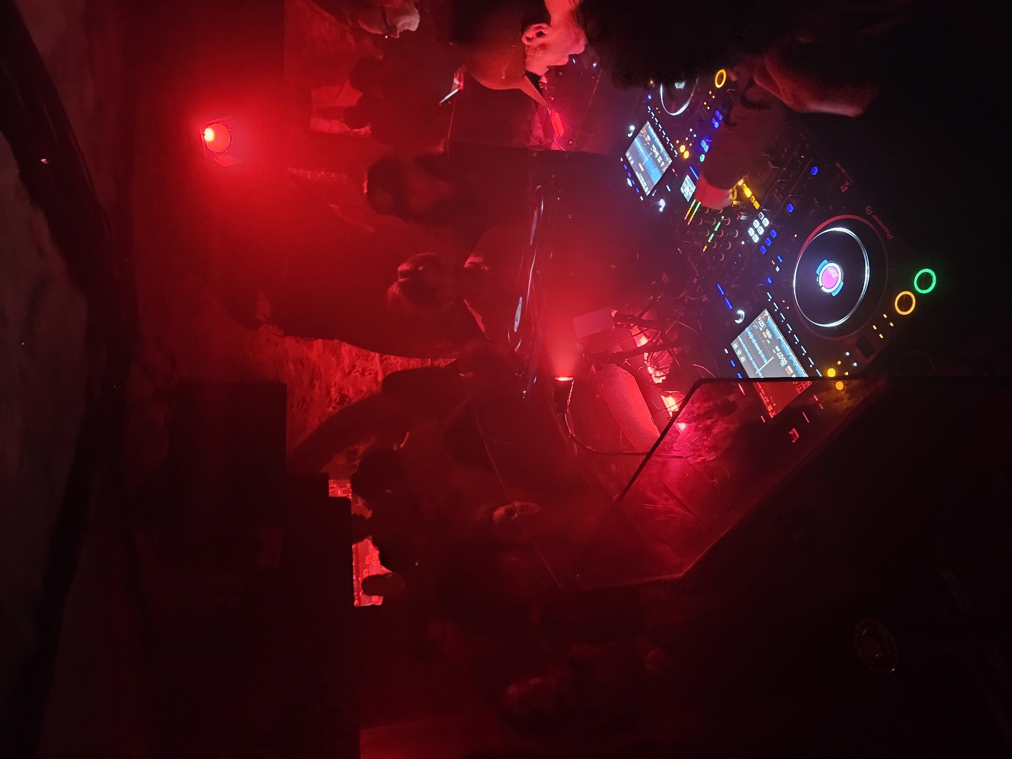 The image captures a DJ booth in a dimly lit, possibly smoky, nightclub setting, recognizable by the prominent DJ equipment, notably Pioneer DJ mixers and turntables with illuminated controls. A group of people surrounds the DJ booth, suggesting a lively atmosphere. The dominant red lighting creates a sense of energy and obscures details. Location cannot be determined from the image alone.