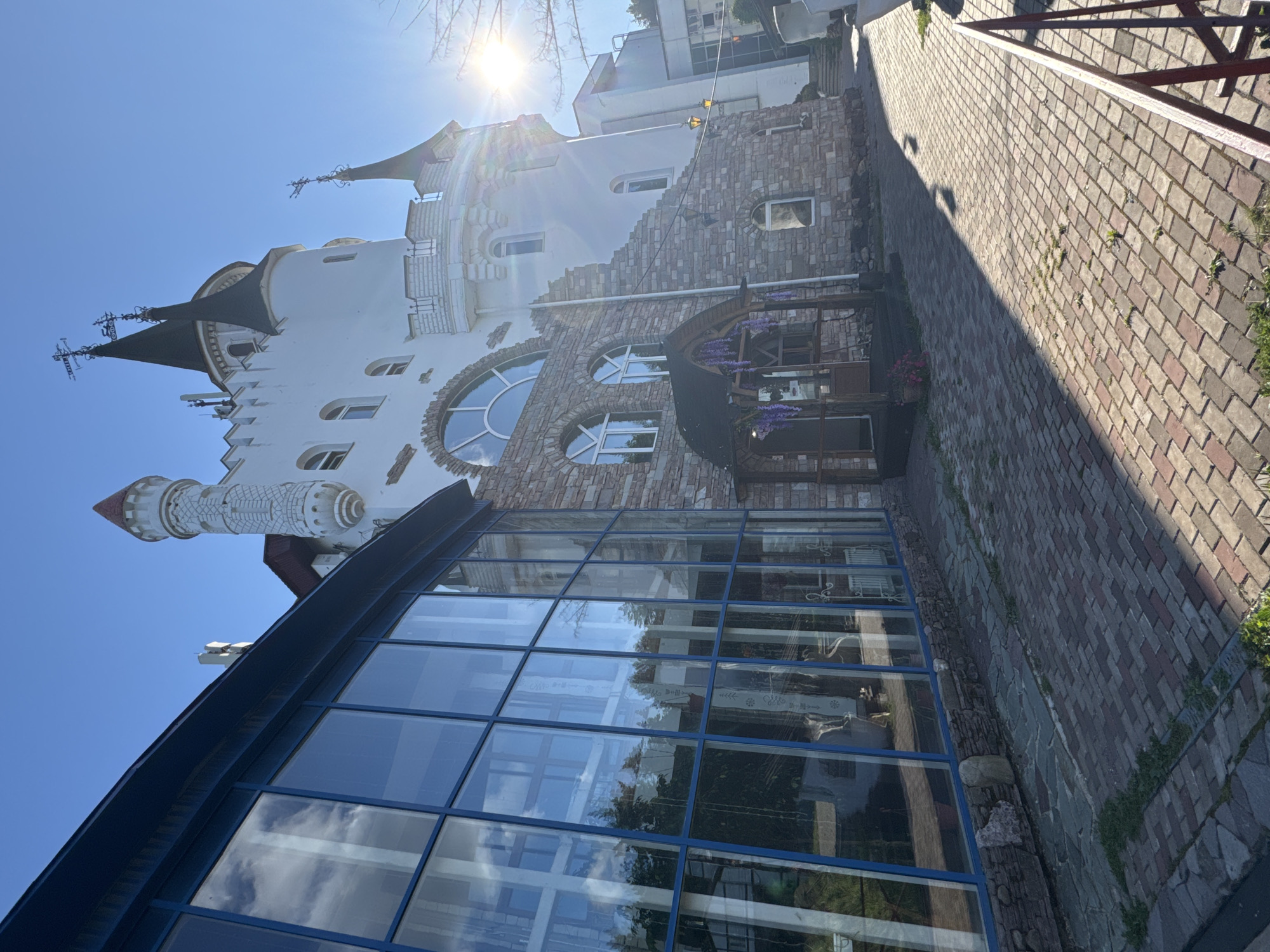 The image shows a building with a whimsical, castle-like design, located in Ufa, Russia. A large, modern glass-walled section with a blue frame is attached to the main building, which features stone accents and white stucco. The castle-like structure has several towers with pointed, dark roofs and decorative elements. The sun is shining brightly, casting long shadows on the brick-paved walkway in front of the building. There are no people visible in the image. The sky is clear and blue. The building appears to be an entertainment venue or a unique architectural attraction.