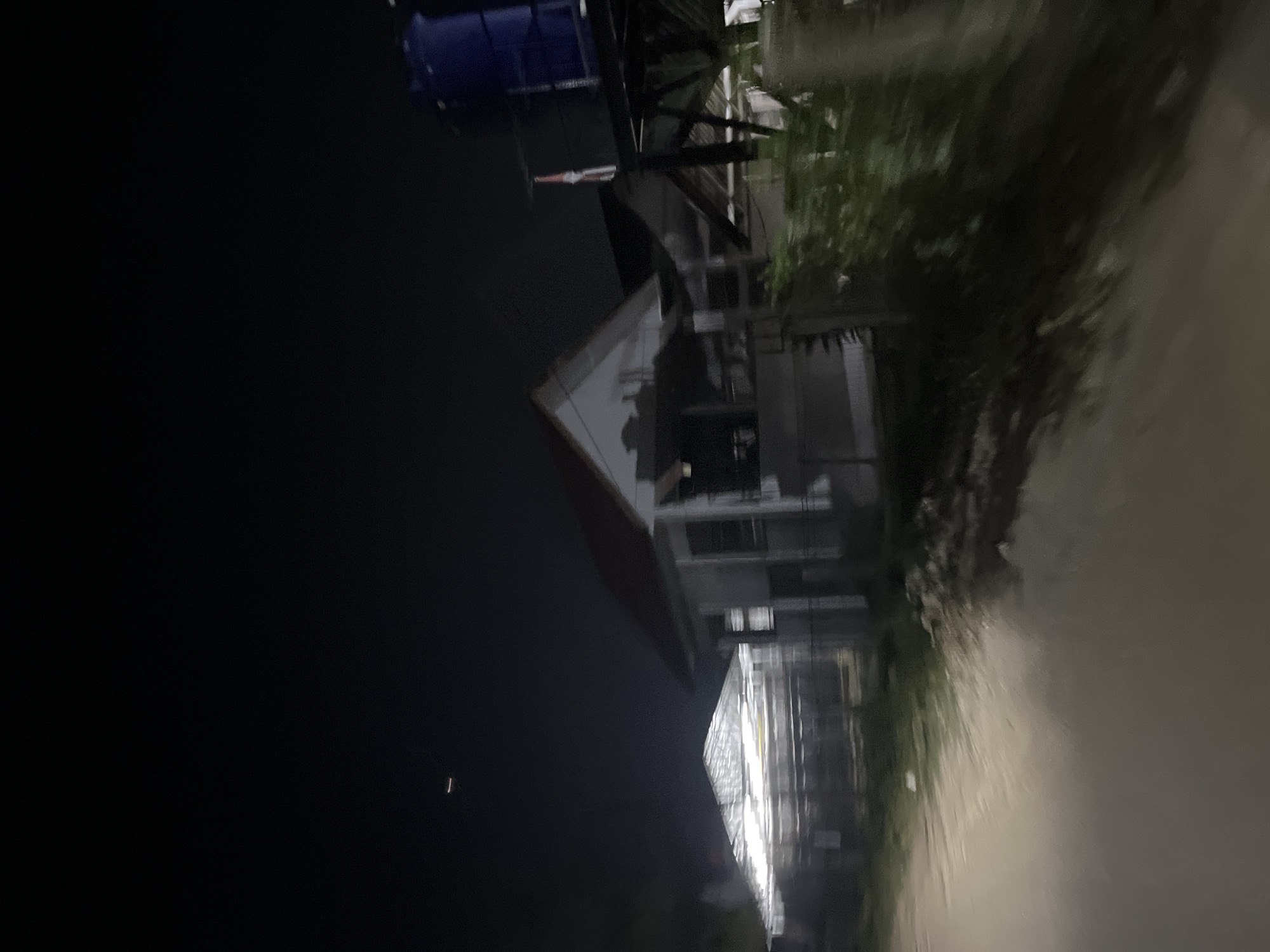 The media file, taken at night in Kesehatanbandar, Indonesia, depicts a dimly lit scene with a focus on a building and its surroundings. A prominent structure, possibly a house or a commercial building, is visible with a distinct triangular roofline and windows. Part of a blue water tank can be seen on top of another structure in the background, suggesting residential or agricultural use. The foreground is dominated by a blurred, light-colored surface, possibly a wet road or a reflective surface, indicating recent rain or high humidity. Lush green vegetation is present, partially obscuring some parts of the building. The overall atmosphere is dark and somewhat hazy, consistent with nighttime conditions. There are no people or specific events clearly captured. The image appears to be taken from a slightly elevated position, looking down on the scene. The lighting suggests artificial illumination from within the building and possibly from streetlights, casting reflections on the wet surfaces.