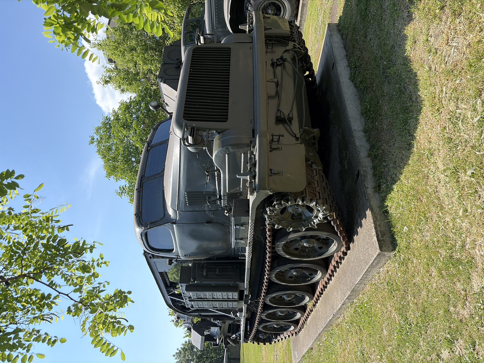 A weathered, olive-drab tracked vehicle, possibly a former military tractor or heavy equipment transporter, is displayed outdoors on a concrete plinth. The vehicle features a prominent grille at the front, a multi-pane windshield, and what appears to be a large crane or lifting mechanism extending from its rear. It is parked on a grassy area with several trees and a bright blue sky with scattered clouds overhead. The vehicle is in a static display, suggesting it is part of a collection or memorial. The location is identified as the National Museum of Military History in Sofia, Bulgaria, a setting that aligns with the presence of historical military artifacts.