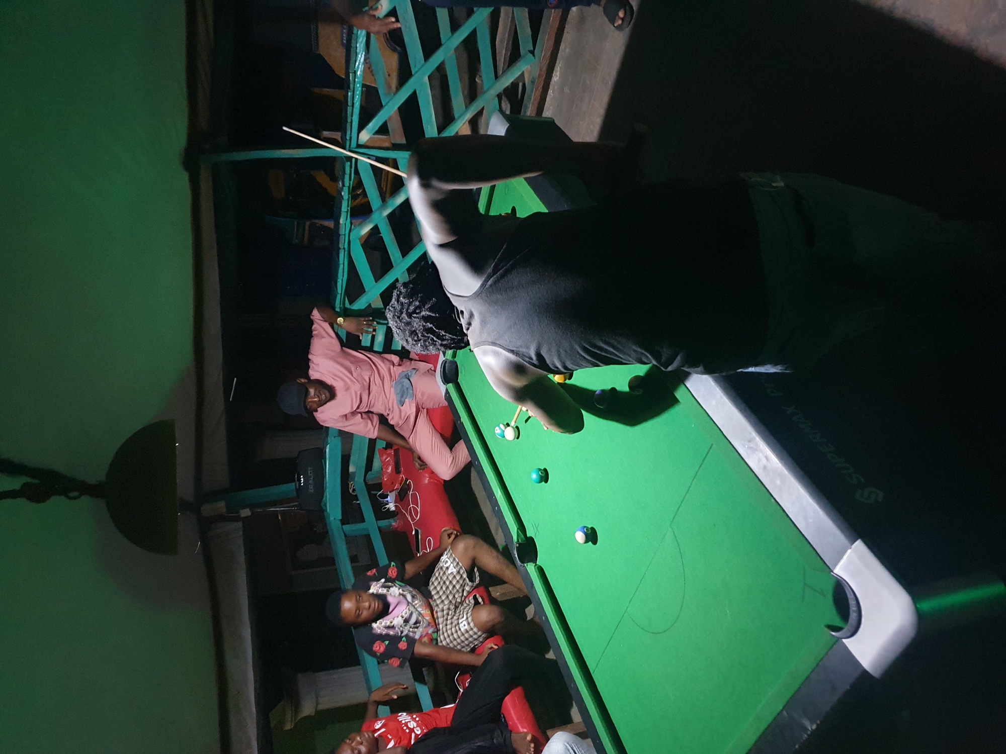 In a dimly lit, likely indoor setting in Lokoja, Nigeria, a group of at least four individuals are present around a pool table. One man, facing away from the camera, is positioned over the table, cue in hand, seemingly taking a shot. Three other individuals are seated along the side of the pool table, appearing to be observing or waiting their turn. The pool table is green with white trim. The lighting casts shadows across the scene, making precise details of the environment difficult to discern.