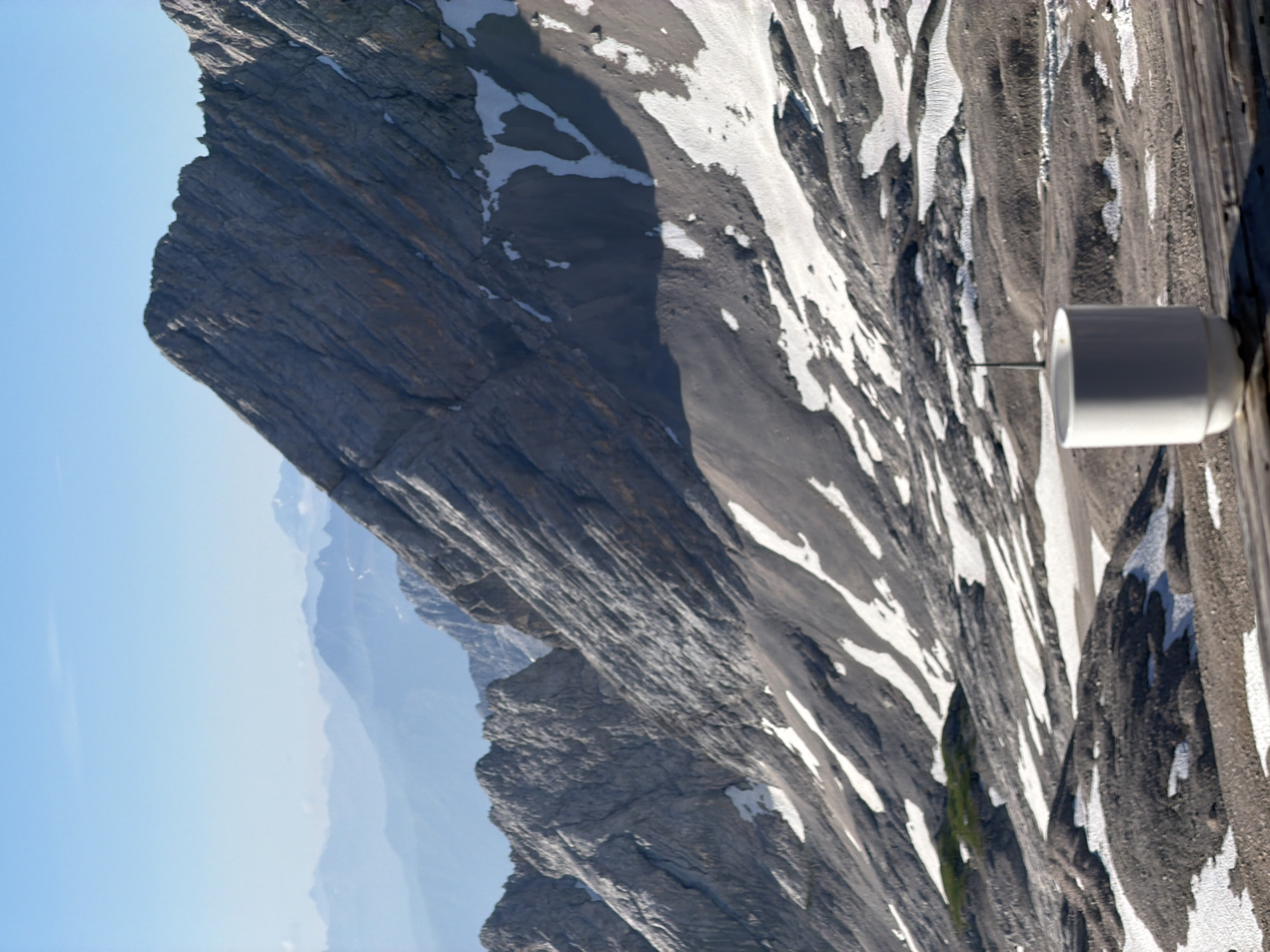 A white ceramic mug sits on a wooden surface in the foreground, with a mountain range in the background. The mug is cylindrical with a slightly tapered base. The mountains are rocky and rugged, with patches of snow. The sky is clear and blue. This image was taken from the Zugspitze, the highest peak in Germany, located near Garmisch-Partenkirchen.