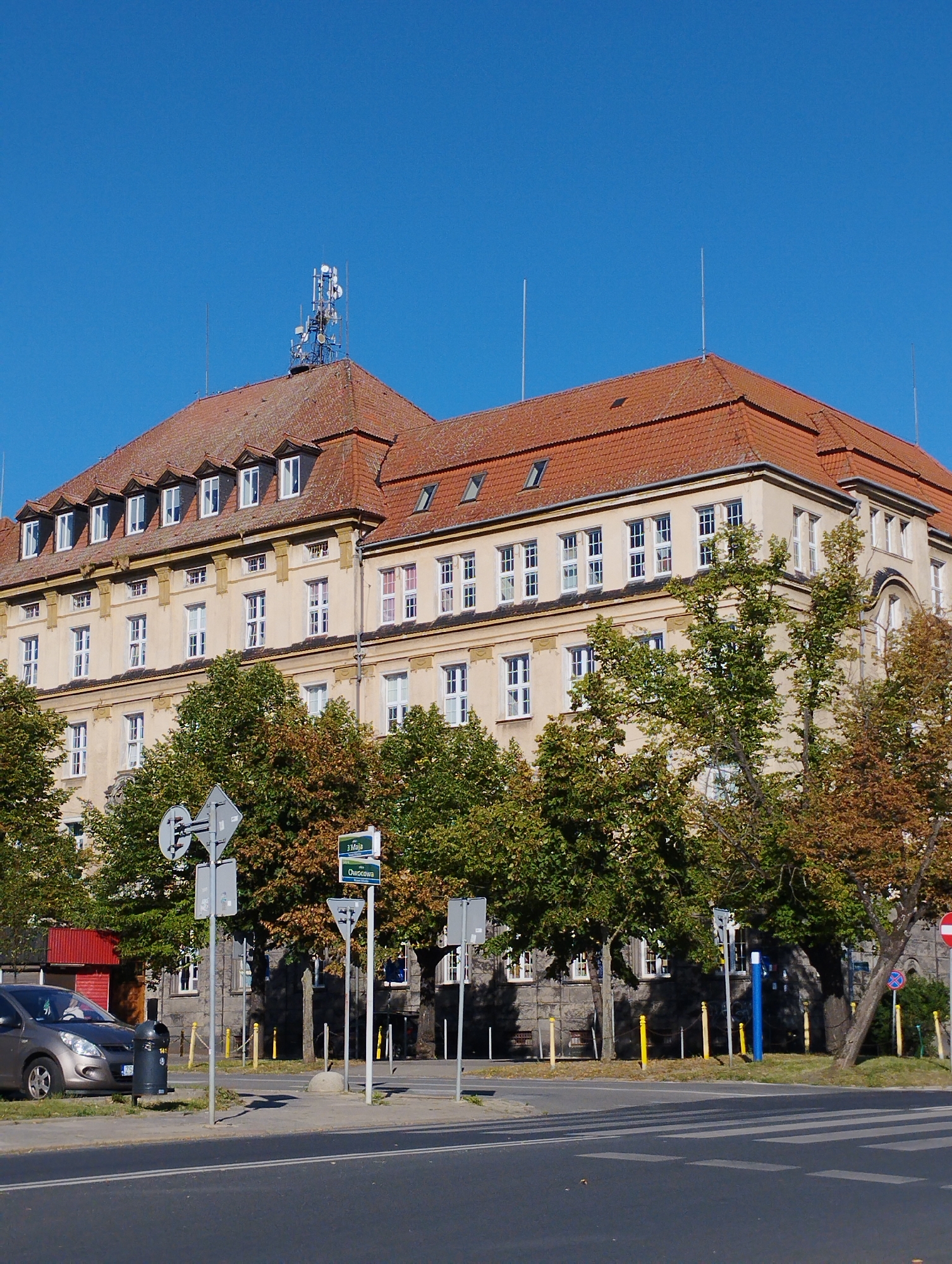 The image shows a multi-story building with a red tile roof, a beige facade, and numerous windows. On the roof, there is a telecommunications tower with multiple antennas. The building is flanked by trees, some of which have autumn foliage. In the foreground, there is a street with a pedestrian crossing and a gray car parked on the side. Several street signs and traffic signals are also visible. The sky is clear and blue. The building is located on ulica Kwiatowa in Szczecin, Poland.