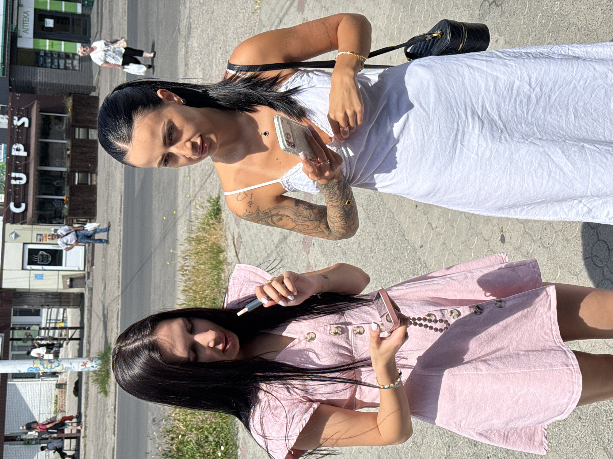 Two women are standing on a sidewalk in front of buildings in an urban environment. The woman on the left has long dark hair and is wearing a pink dress. She is holding a vape pen in her right hand and a small rose-gold colored case in her left hand. The woman on the right has dark hair pulled back in a ponytail and is wearing a white dress. She is holding a smartphone in her right hand and has a black purse slung over her left shoulder. In the background, there are several people walking and standing, and shops are visible with signage that reads "CUPS" and "АПТЕКА". The ground is paved with cobblestone-like patterns, and there is a street with a sidewalk bordering it.