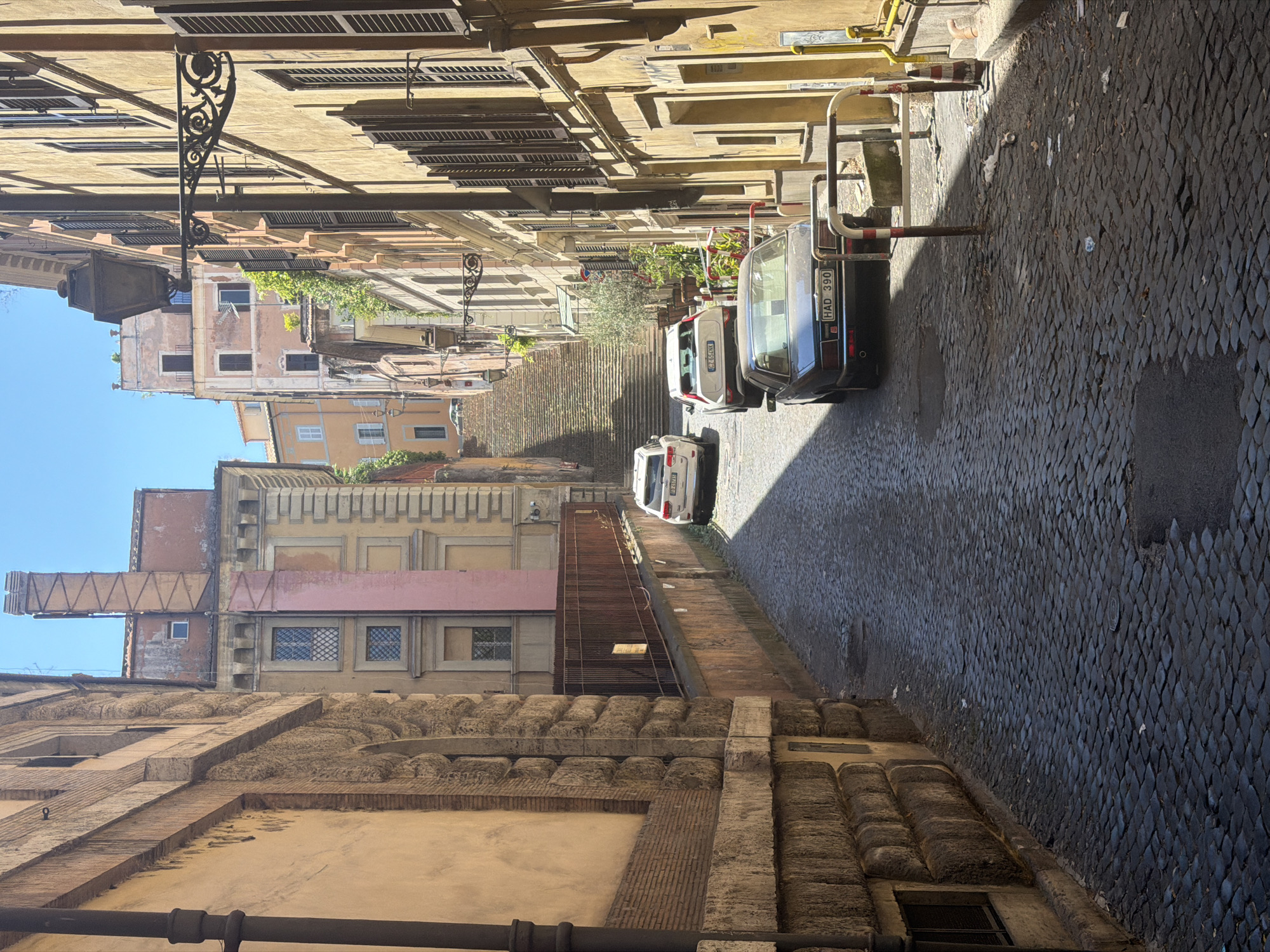 In a narrow, cobblestone street in an old city, possibly Rome, Italy, several cars are parked and driving. The street is lined with tall, old buildings on both sides, featuring weathered facades, numerous windows with dark shutters, and architectural details like balconies and cornices. On the left, a large, stone building with ornate carvings dominates the foreground. On the right, a building with a yellow facade and dark shutters casts a shadow across the street.
In the distance, a long, wide staircase ascends, leading to a higher part of the city, with more buildings visible beyond. The sky is clear and blue. The scene is bathed in strong sunlight, creating distinct shadows. The cobblestone street is uneven, with some areas appearing wet or darker than others. Litter is visible on the ground in the lower right section of the image.
Parked on the street are three cars: a white SUV in the middle distance, a white hatchback further back, and a dark-colored sedan in the foreground. A brown metal fence runs along the right side of the street for a section. A street lamp with an ornate bracket is attached to the building on the right. The overall impression is of a historic European urban environment, likely during the daytime.