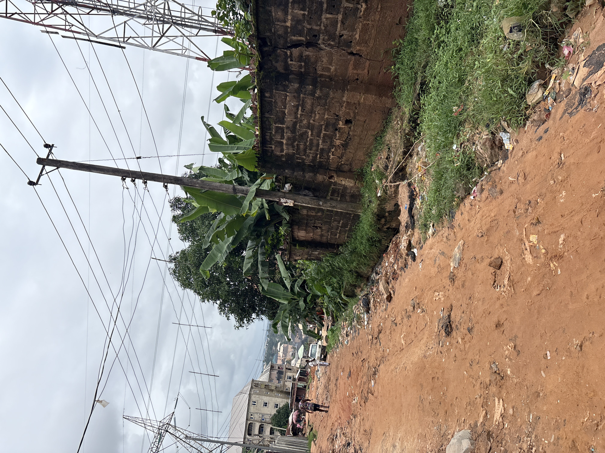 The image depicts a scene on a red dirt road in Nkpor, Nigeria. The road is unpaved and appears to be in disrepair, with visible potholes and debris scattered across its surface. On the right side of the road, a low brick wall runs along the edge, topped with lush green banana plants. Beyond the wall, there is a drainage ditch filled with overgrown grass and a significant amount of litter.
In the foreground, two individuals are walking away from the camera on the dirt road. One person, a male wearing a patterned shirt and shorts, is in the process of walking. Another person, dressed in white clothing, is walking beside a white vehicle parked on the road. In the background, a row of multi-story buildings lines the left side of the road, with some appearing to be shops or residences. Overhead, a complex network of power lines crisscrosses the sky, supported by wooden utility poles and a tall metal transmission tower visible on the right edge of the frame. The sky is overcast, suggesting cloudy weather. The overall impression is one of a working-class urban environment with infrastructure challenges and visible signs of litter.