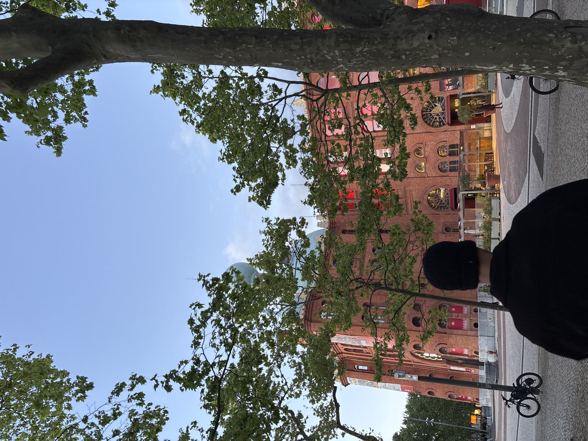 A low-angle shot looking up at the Campo Pequeno bullring in Lisbon, Portugal. A person wearing a black knit cap and black jacket stands on the sidewalk. A light pole with a bicycle locked to it are near the person. Branches and foliage from a large tree frame the top of the image, while the bullring dominates the middle. The building is red brick with decorative windows. The sky is light blue.
