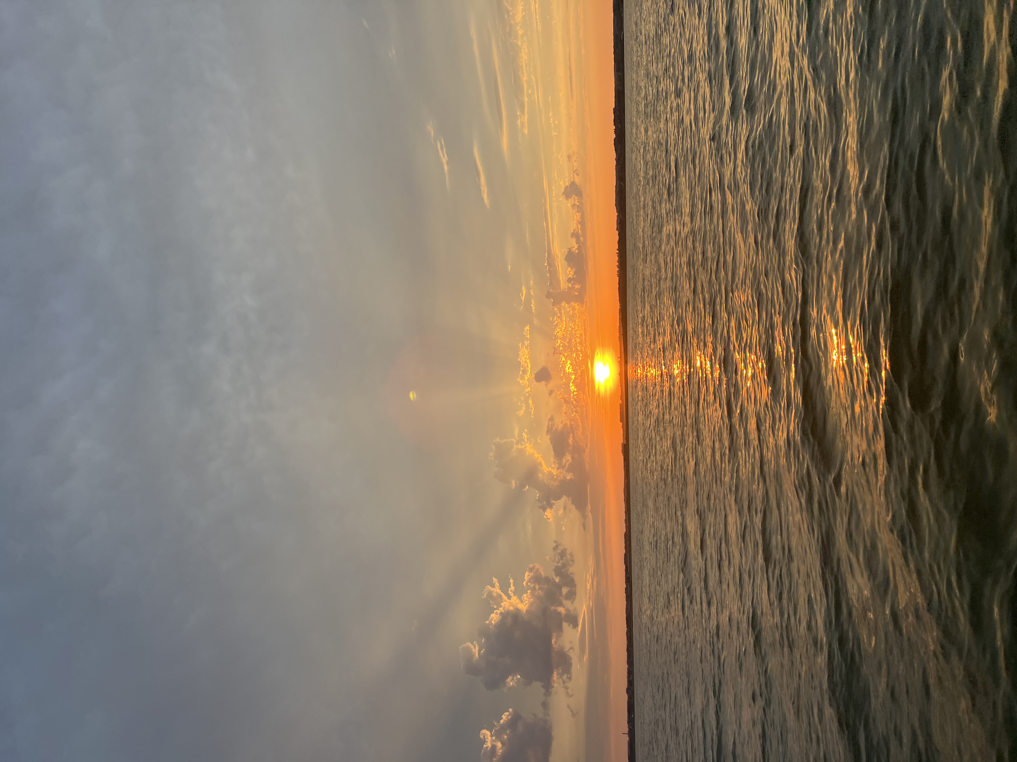 The image depicts a serene sunset over a body of water. The sky is a gradient of warm orange and yellow near the horizon, transitioning to muted grey and blue clouds higher up. Rays of sunlight emanate from the setting sun, which is partially obscured by clouds. The water is calm, with gentle ripples reflecting the colors of the sky, creating a shimmering effect. A faint, distant shoreline with structures is visible against the horizon. The scene is tranquil and devoid of any human presence. The location is identified as Novopichugovo, Russia.
