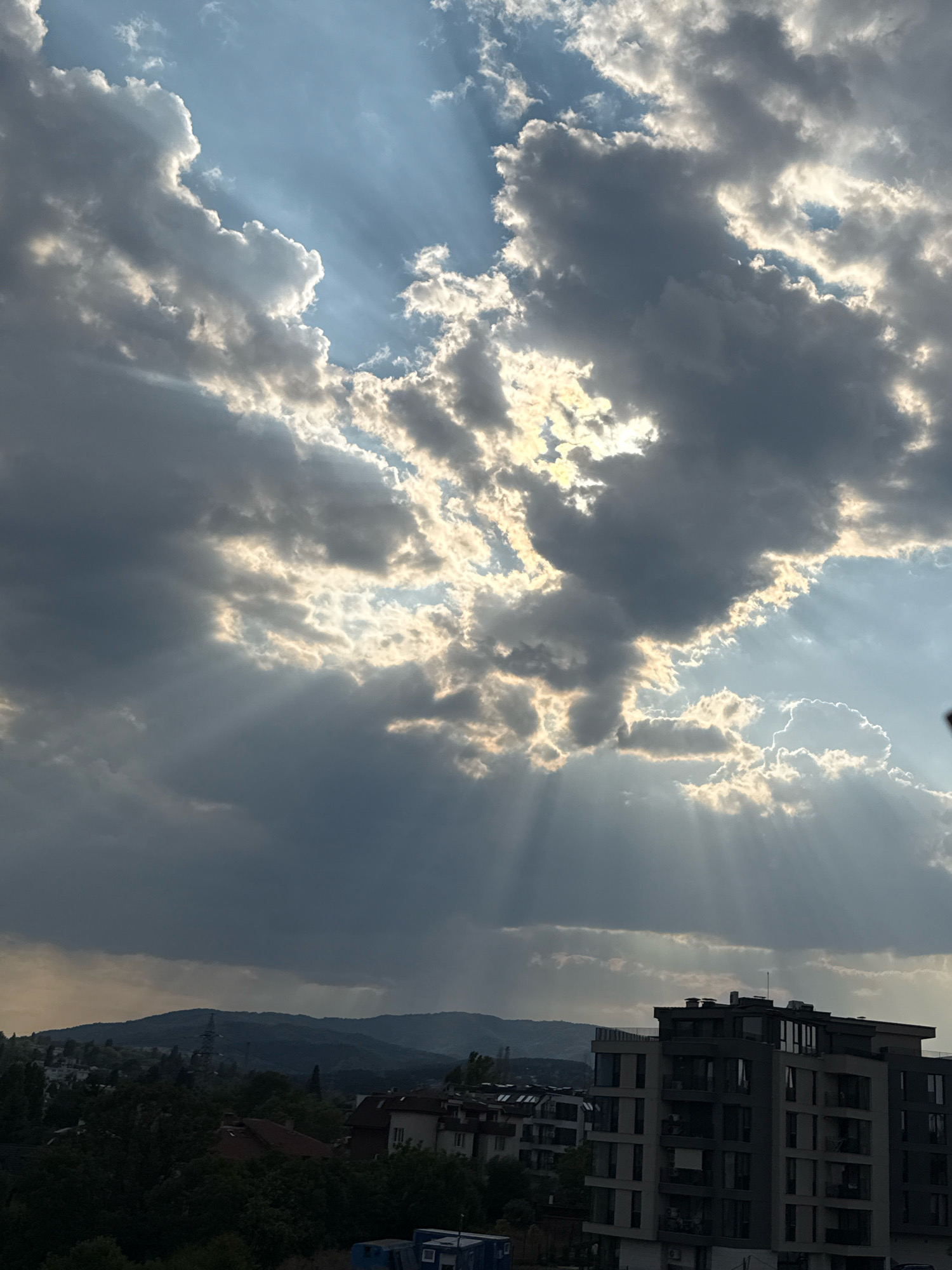 This is a medium-shot, vertical, eye-level photograph taken from a high vantage point overlooking a city with a mountainous backdrop. The sky dominates the upper portion of the frame, filled with dramatic, scattered clouds illuminated by crepuscular rays from the sun. The clouds are a mix of dark grey and bright white, with streaks of sunlight breaking through in various parts of the sky.
Below the sky, the middle ground shows a dense collection of trees and buildings, suggesting a densely populated urban area. The buildings are of various heights and architectural styles, with some appearing to be residential apartments and others possibly commercial structures. The closest building on the right is a modern, multi-story apartment building with a grey facade, balconies, and numerous windows. Further in the distance, a range of hazy mountains forms the horizon, partially obscured by atmospheric conditions. The lower part of the image is dark, possibly due to shadows cast by the clouds or the time of day.
The scene depicts a cityscape during daytime, likely in the late afternoon or early evening given the dramatic lighting from the sun breaking through the clouds. The environment is urban, with a clear presence of both natural elements (sky, clouds, trees, mountains) and man-made structures (buildings). There are no discernible people or active interactions visible in the frame. The overall mood is one of natural beauty and urban coexistence. The presence of the specific modern apartment building on the right, the dense urban environment, and the mountainous horizon are characteristic of certain neighborhoods in Sofia, Bulgaria, particularly those on the outskirts closer to the Vitosha Mountain range, such as the Lozenets or Studentski Grad districts.