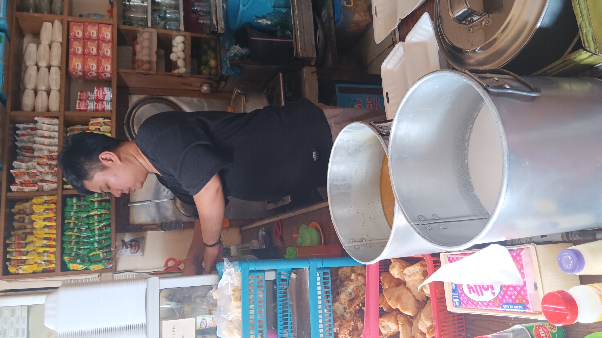 The image depicts the interior of a small food stall or eatery, likely in the city of South Jakarta, Indonesia. A man, appearing to be of East Asian descent, with short black hair and a dark t-shirt, stands behind a counter. He is in the process of preparing food or serving customers, with his hands engaged in an activity.
The stall is densely packed with various items. Shelves lining the back wall are filled with numerous packaged food products, including instant noodles in yellow and green packaging, as well as what appear to be boxes of biscuits or snacks. Various containers, including cans and jars, are also visible on the shelves, alongside what look like eggs and citrus fruits.
In the foreground, there are several large aluminum containers, one filled with a yellowish liquid and another with a white liquid. There are also plastic containers holding fried food items. Several bottles of condiments and sauces are on the counter, including one with an "ABC" label. A stack of white disposable food containers is also present. The overall impression is of a busy, functional food preparation area. The lighting suggests it is daytime, possibly indoors with artificial lighting.